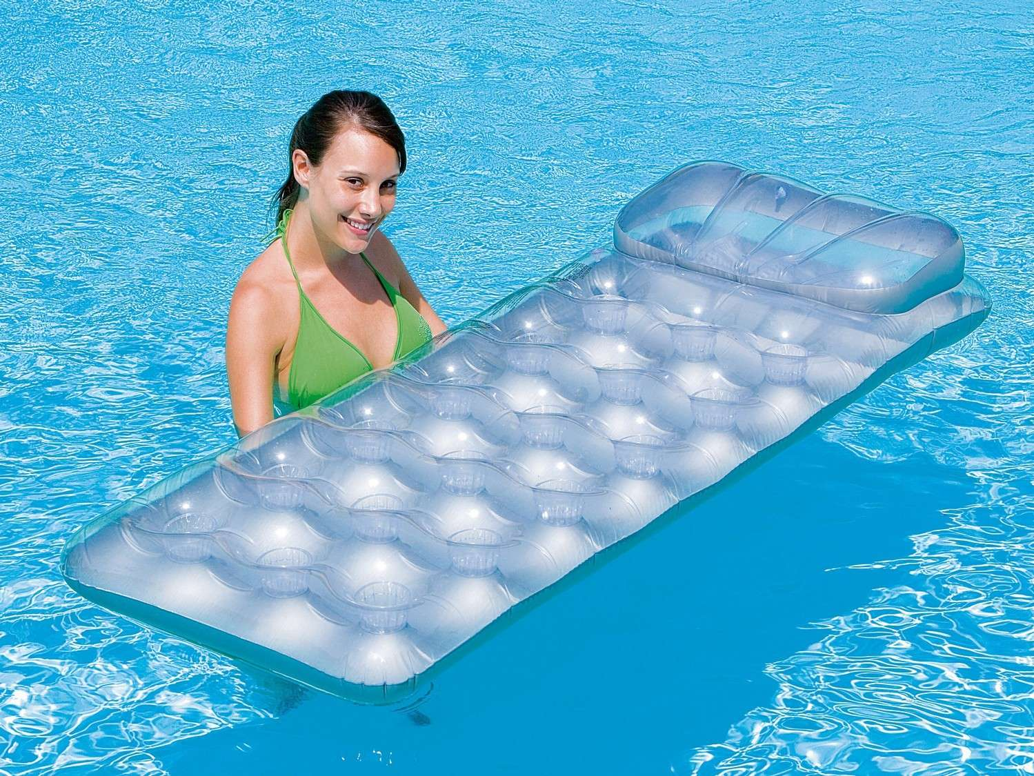 Una mujer en bikini verde sonríe mientras sostiene una balsa inflable transparente en una piscina azul.