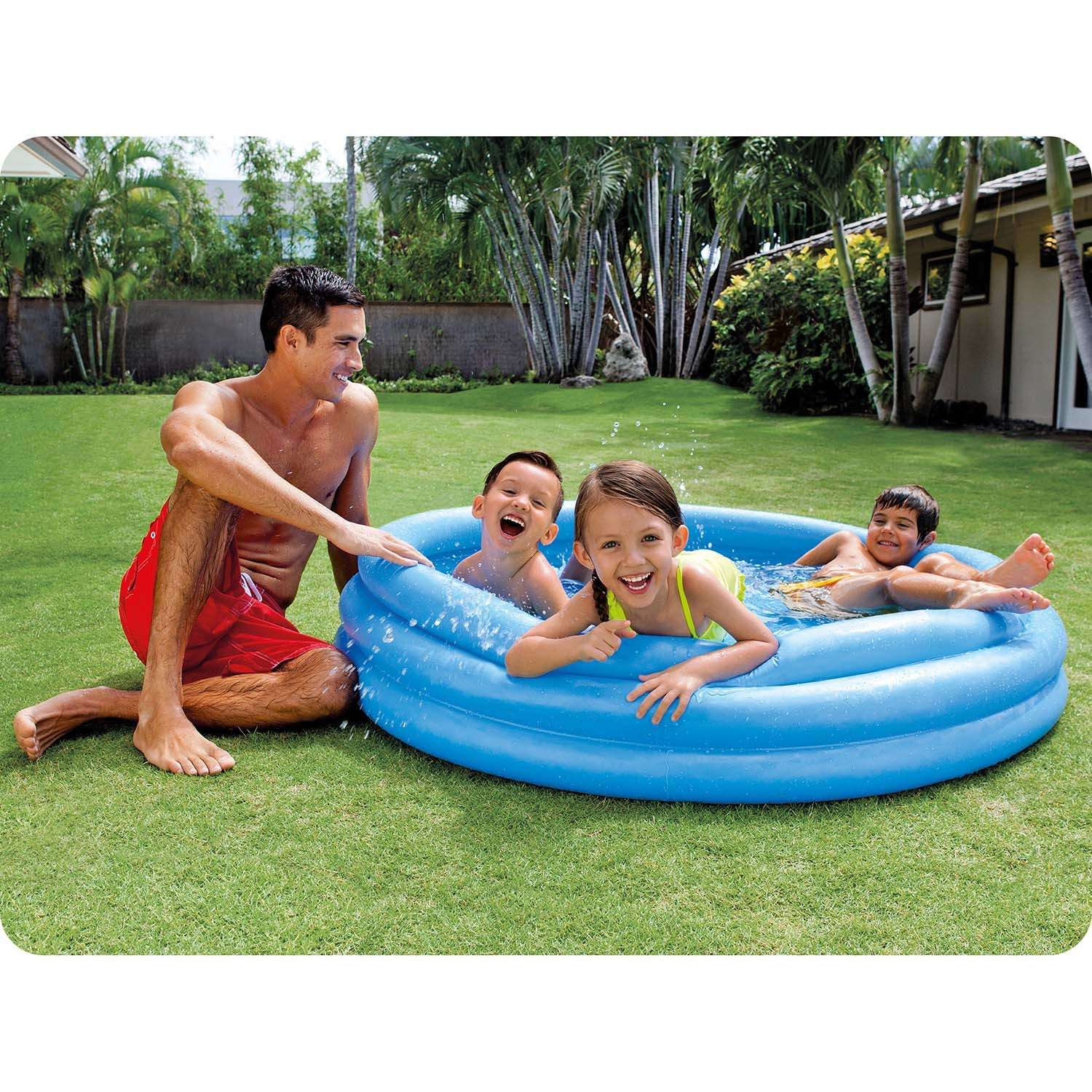 Un hombre y tres niños están en una piscina inflable azul sobre césped verde. Sonríen y se divierten. Hay palmeras al fondo.