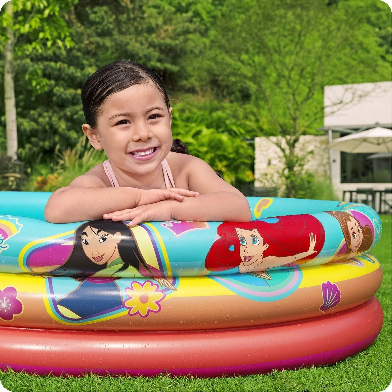 Una niña sonríe en una piscina inflable colorida con diseños de princesas Disney. Hierba verde y árboles en el fondo.