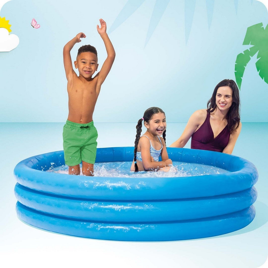 Niños y una mujer en una piscina azul. Un niño se para con los brazos arriba, una niña se sienta, la mujer sonríe. Palmera y sol al fondo.