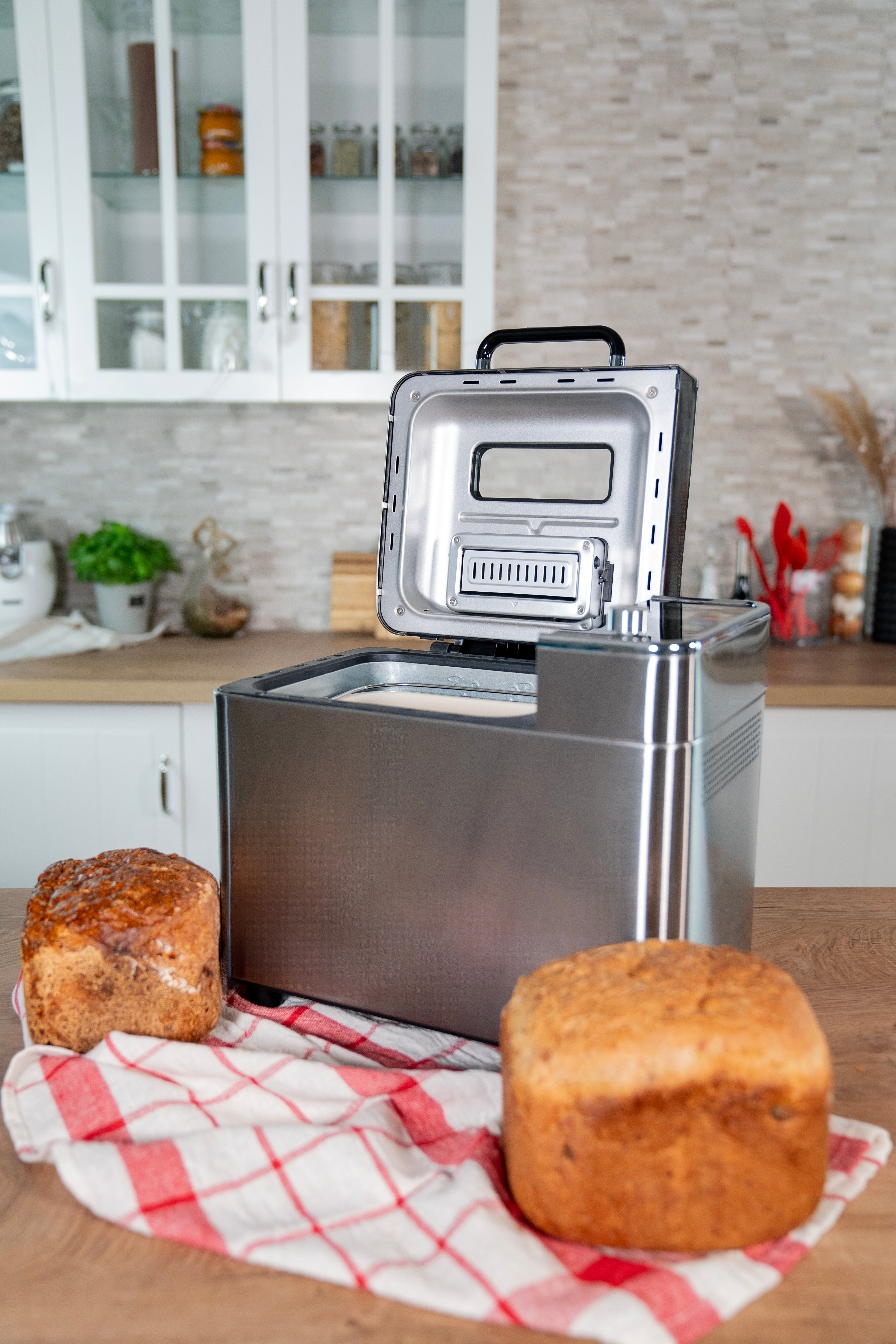 Brotbackautomat mit offenem Deckel und zwei Brotlaiben auf kariertem Tuch. Küchenhintergrund.