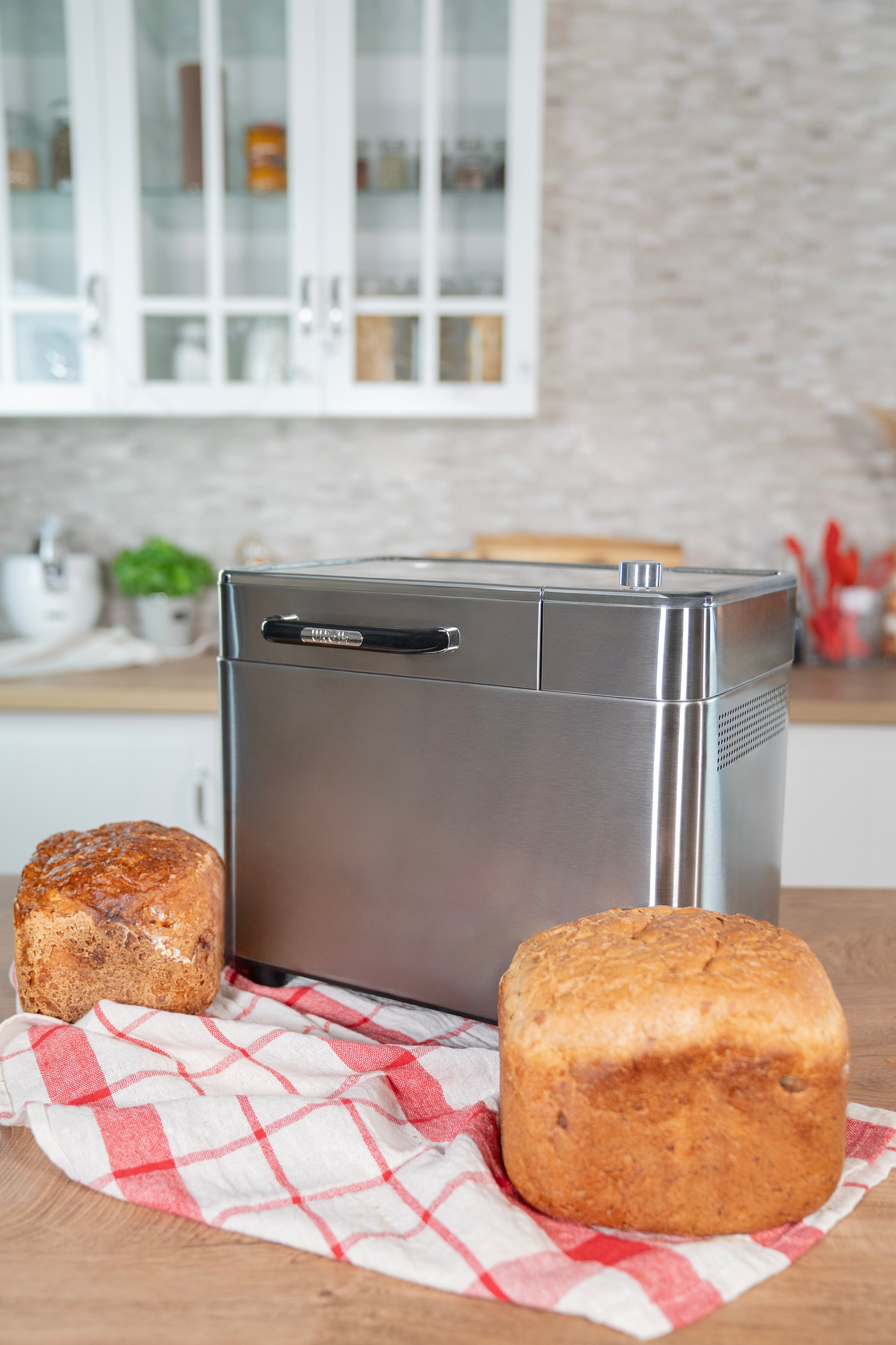 Eine Brotbackmaschine mit zwei Brotlaiben auf einem karierten Tuch. Küchenhintergrund mit Schränken und einer Theke.