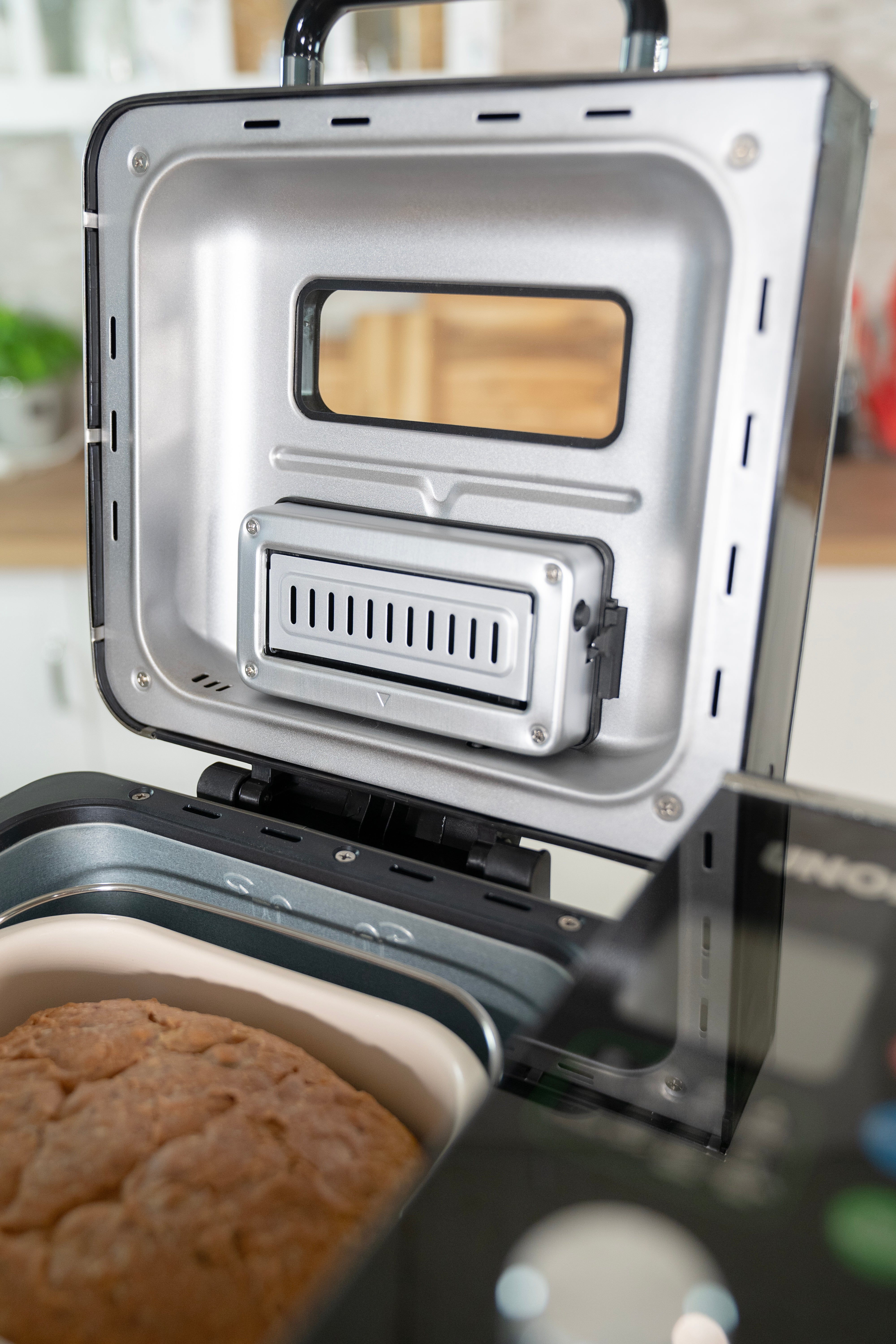 Geöffneter Brotbackautomat mit Brot. Silbernes Innere, schwarzer Griff. Fenster zum Hineinsehen. Küchenhintergrund.