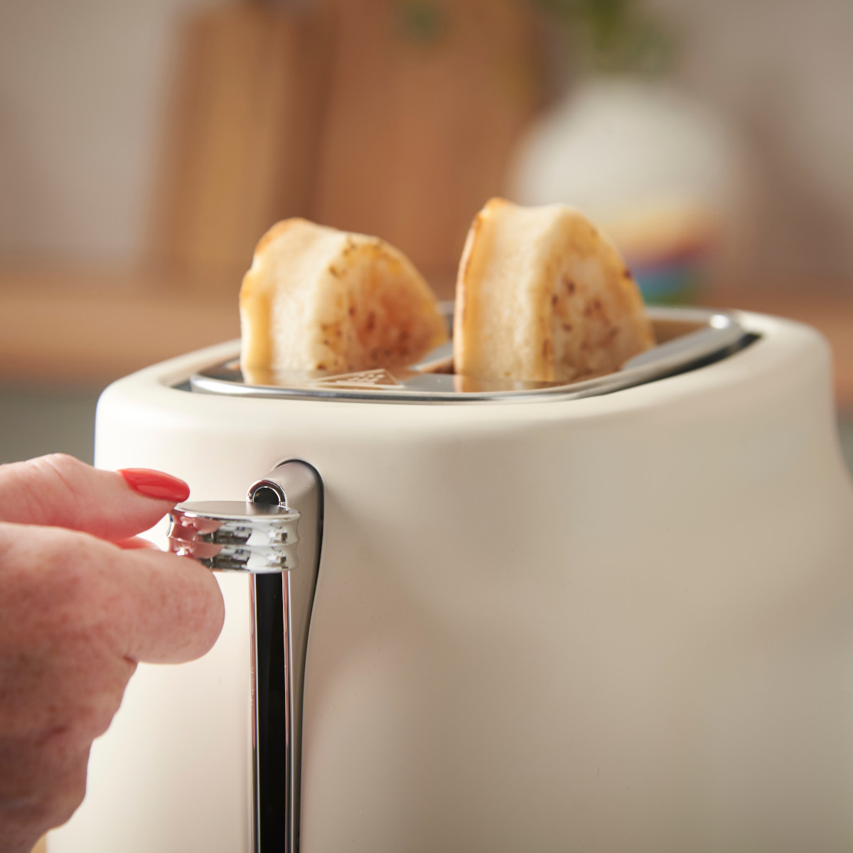 Eine Hand mit roten Nägeln zieht den Hebel an einem cremefarbenen Toaster mit zwei Toastscheiben. Der Hintergrund ist unscharf.