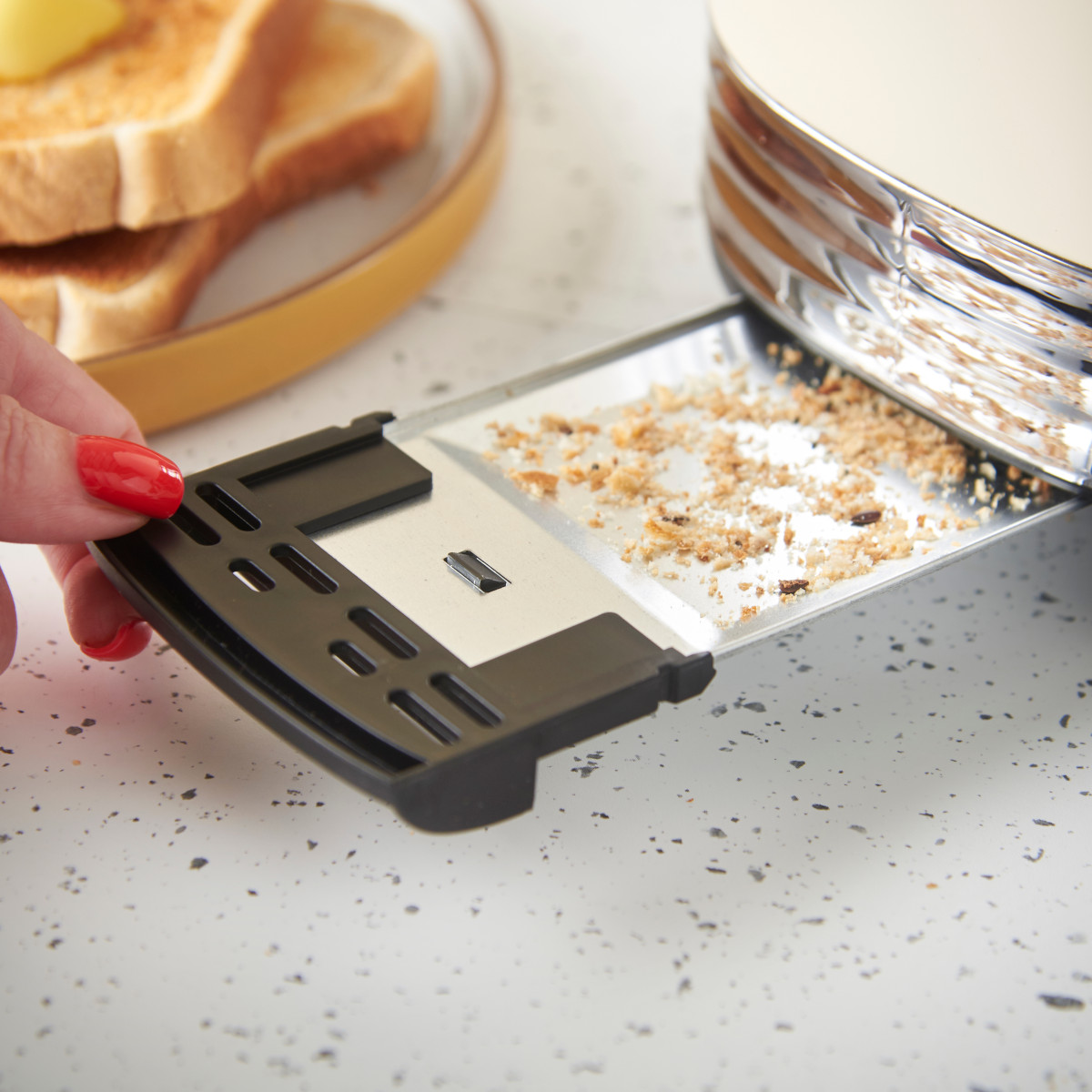 Eine Hand mit roten Nägeln zieht eine Krümelschublade aus einem Toaster. Getoastetes Brot mit Butter liegt auf einem Teller im Hintergrund.