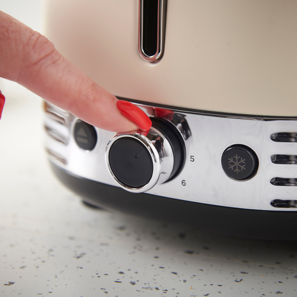 Eine Hand mit rotem Nagellack dreht einen Drehregler an einem cremefarbenen Toaster. Die Zahlen 3, 5 und 6 sind sichtbar, sowie eine Schneeflockentaste.