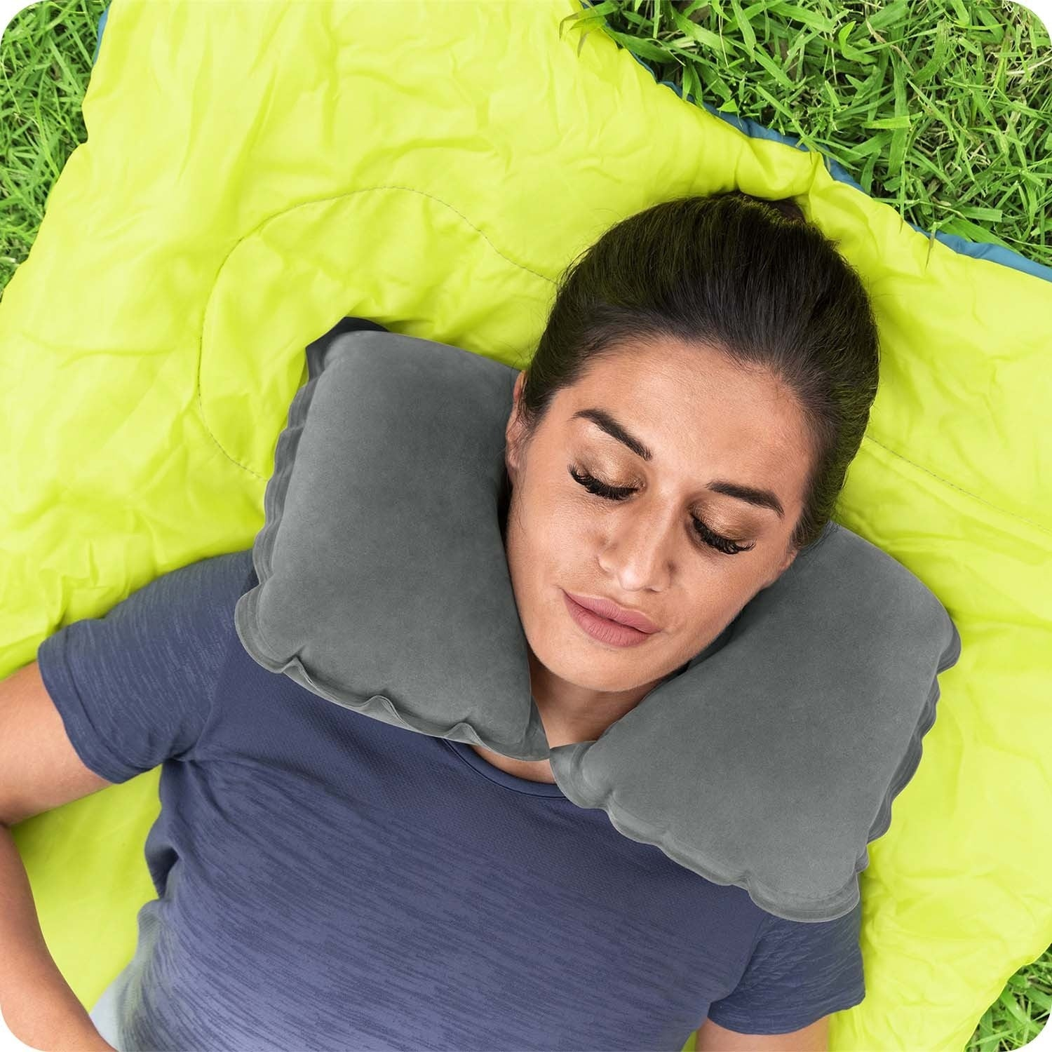 Una mujer descansa al aire libre sobre una colchoneta verde, con una almohada gris para el cuello. Está sobre césped, con una camisa azul, con los ojos cerrados.