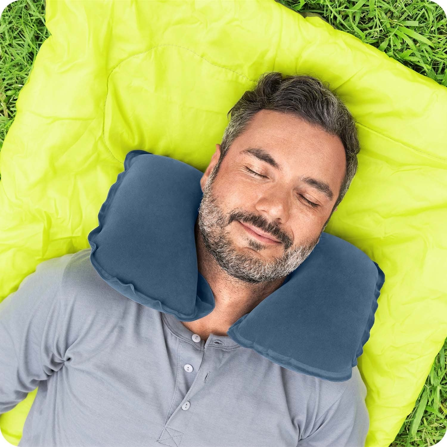Un hombre duerme sobre una manta verde neón en la hierba, con una almohada azul para el cuello. Tiene barba y lleva una camisa gris.