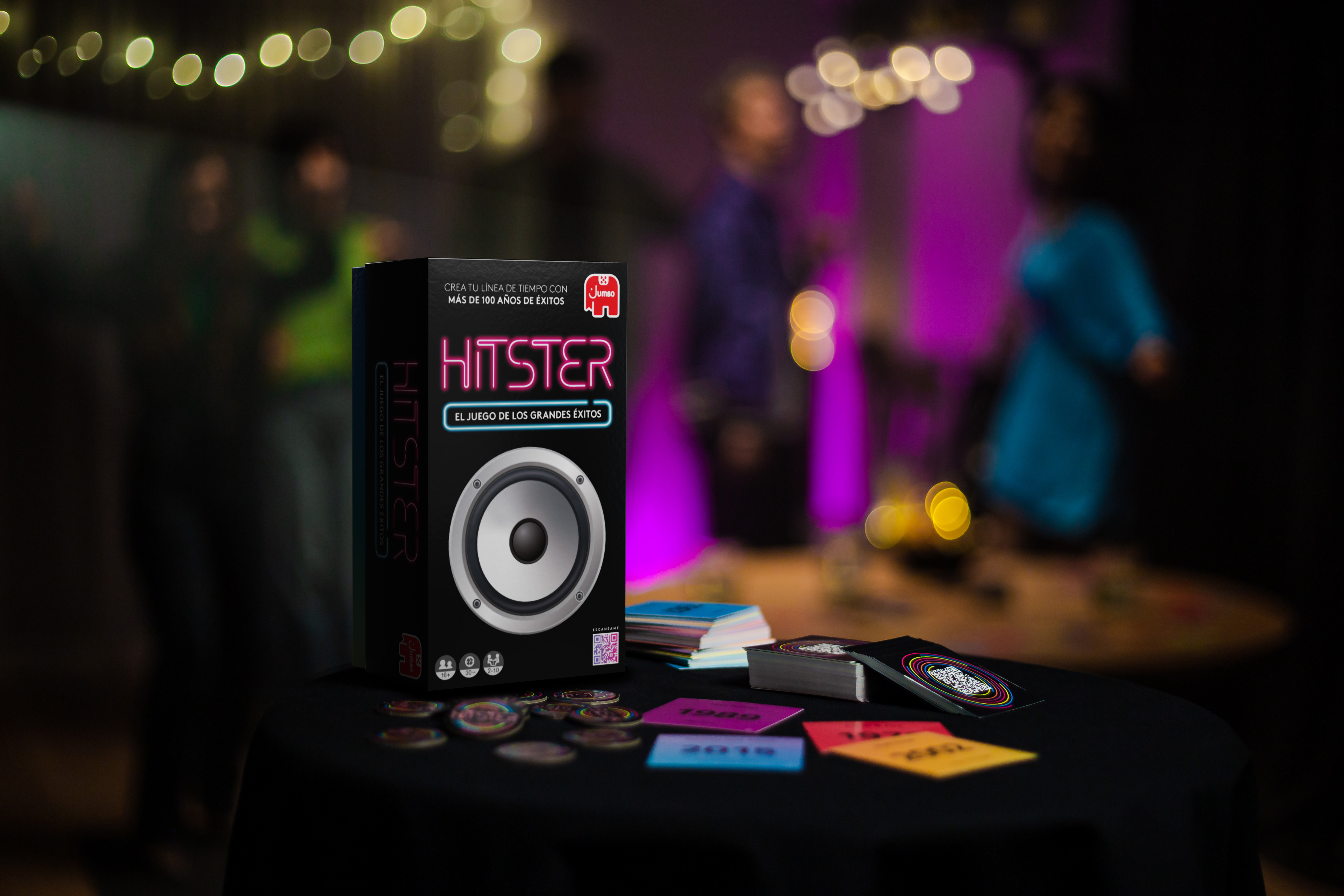 Una caja de juego 'Hitster' está sobre una mesa con cartas. Hay personas borrosas en el fondo, con luces.