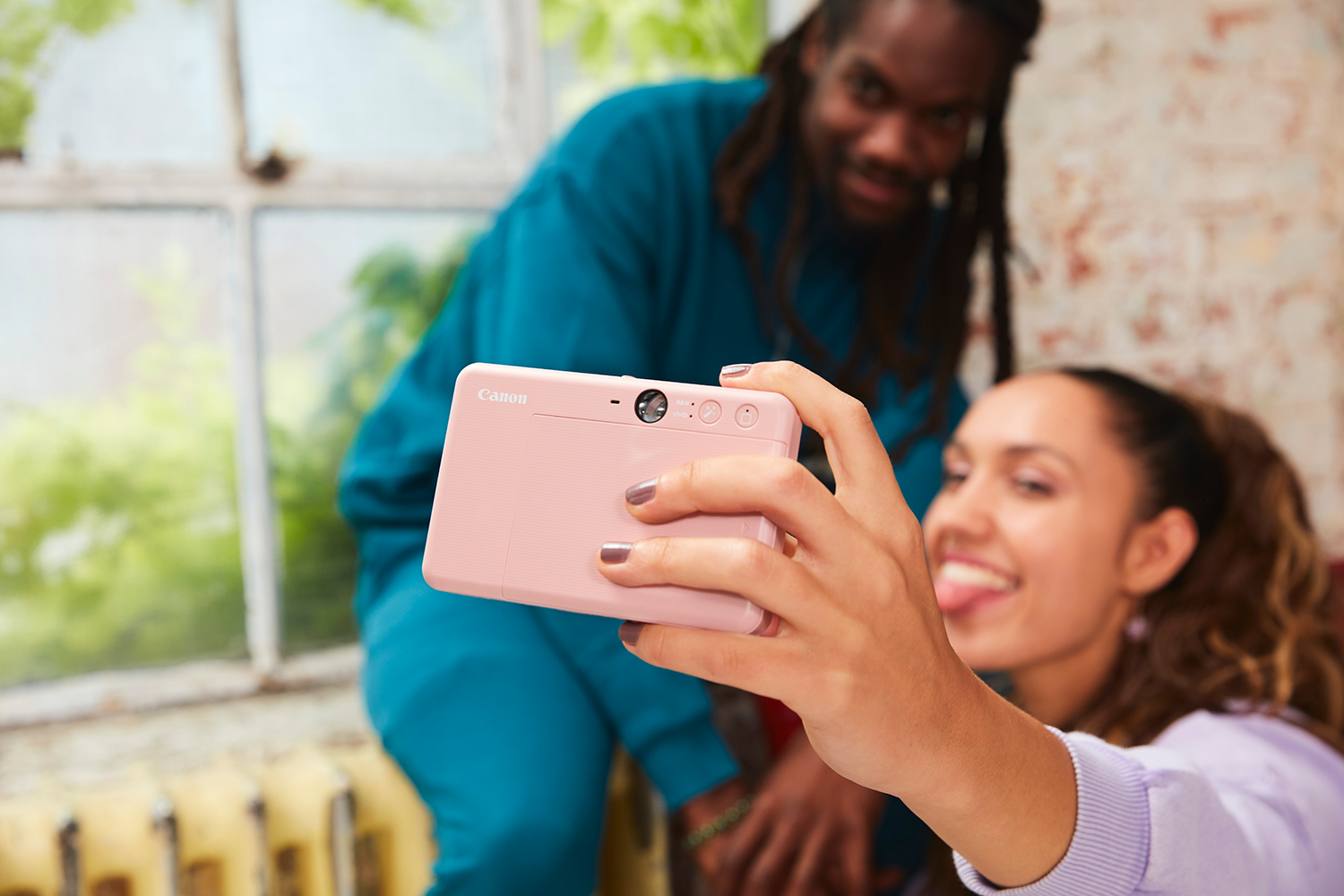 Een vrouw maakt een selfie met een roze camera en steekt haar tong uit. Een man lacht op de achtergrond. De camera is Canon.