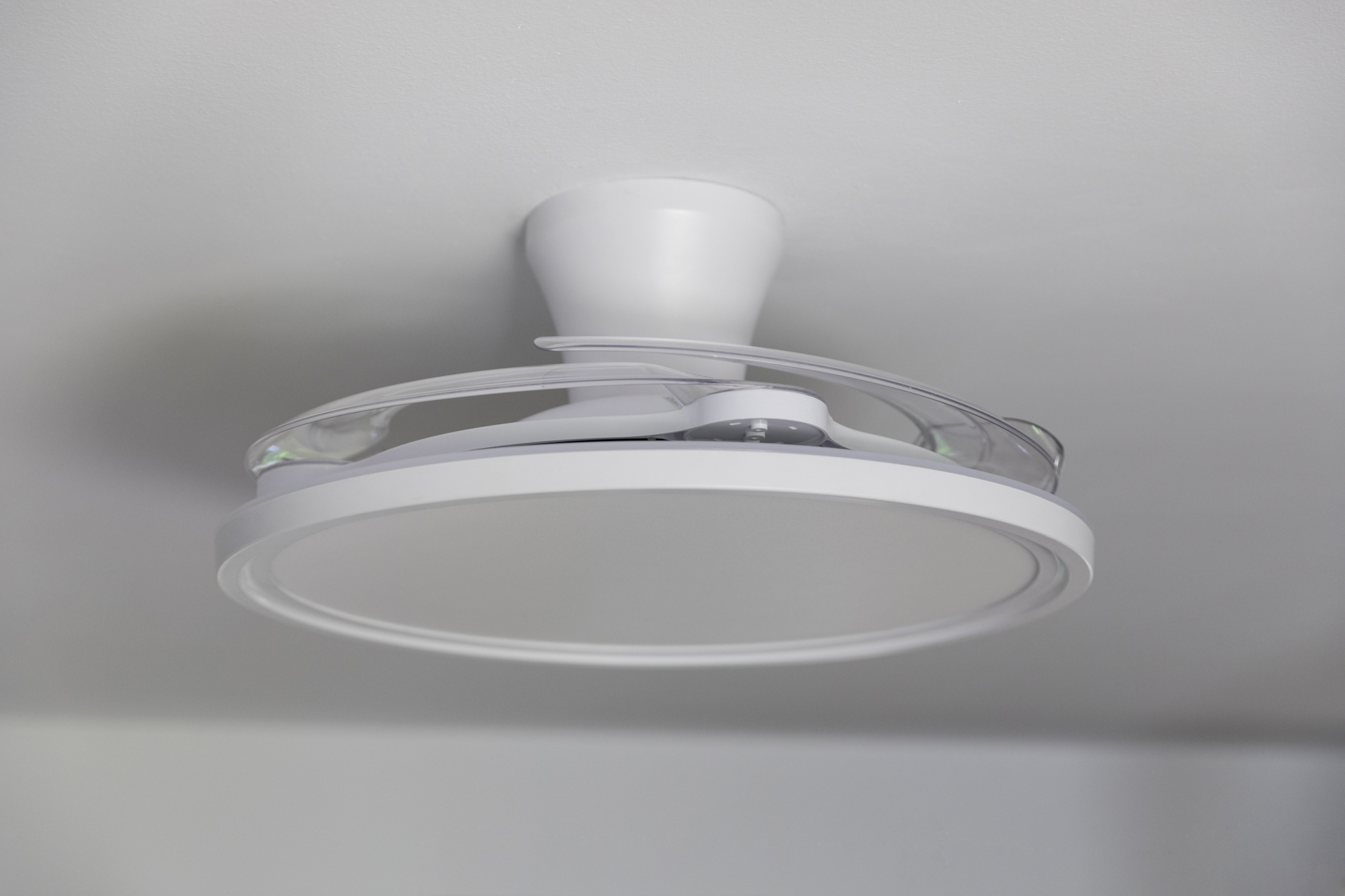 Un ventilador de techo blanco con aspas transparentes y una lámpara redonda cuelga de un techo blanco.
