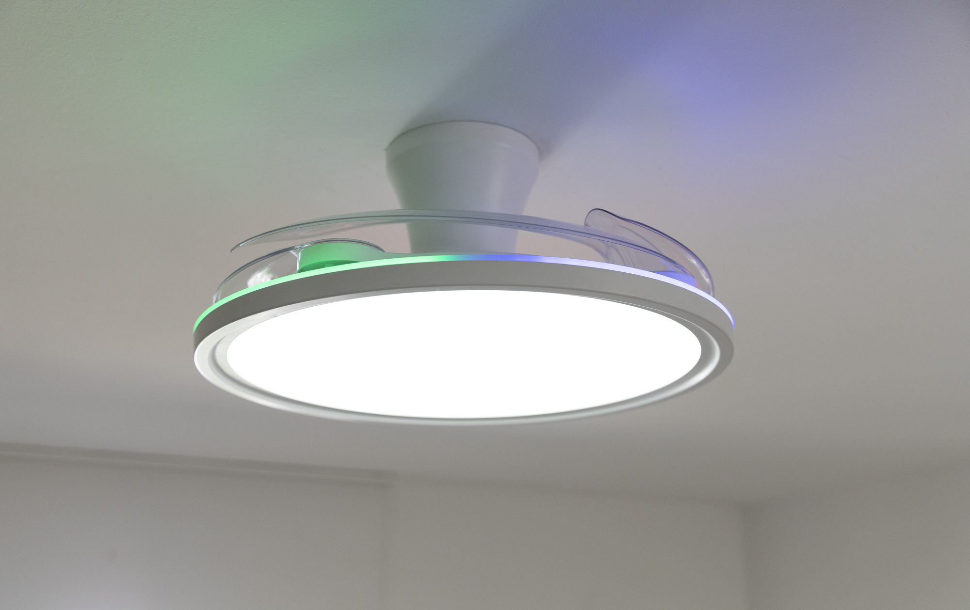 Un ventilador de techo moderno con luz circular brillante. Los bordes tienen iluminación verde y azul. Montado en techo blanco.