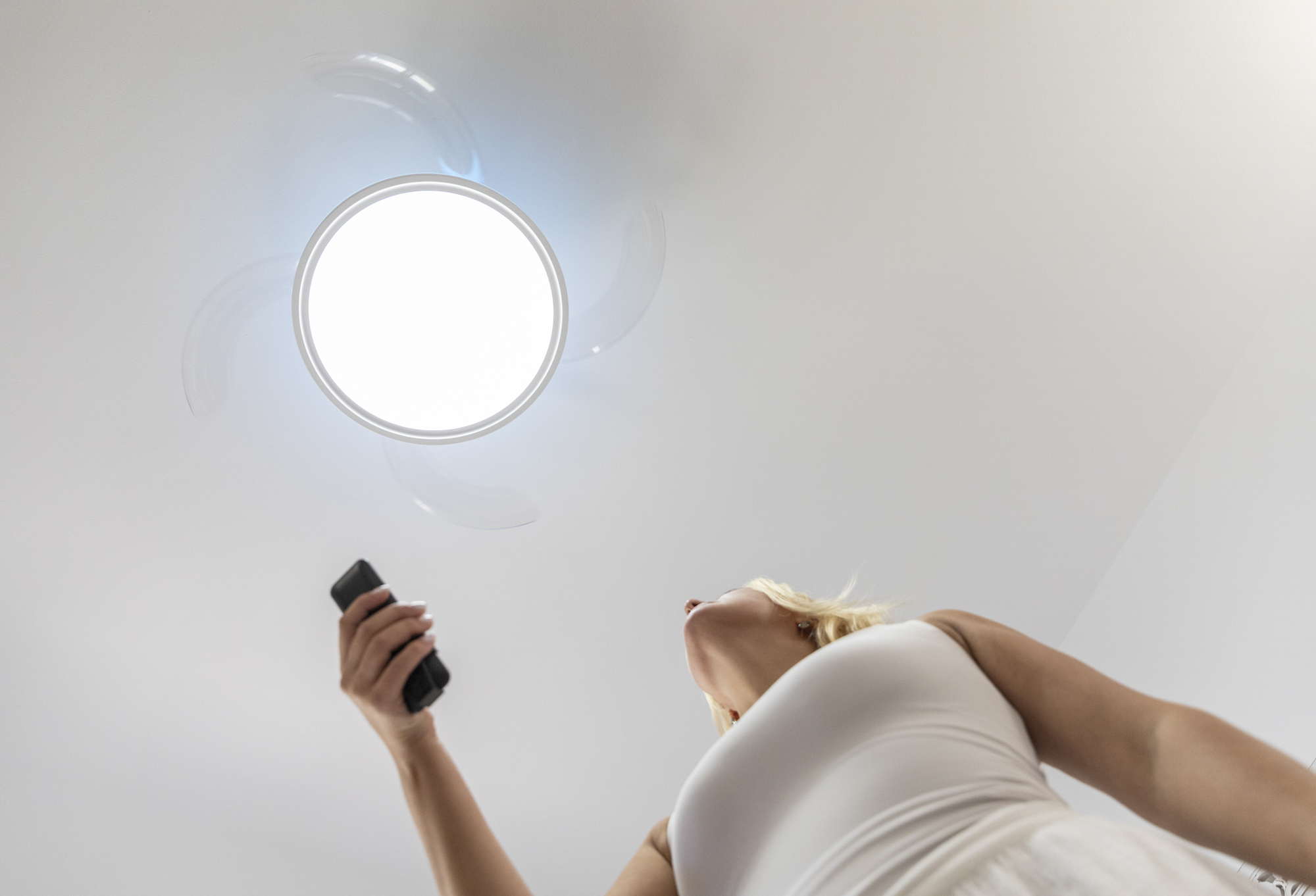 Una mujer con un teléfono mira hacia arriba a una luz de ventilador de techo. Las aspas del ventilador están borrosas. La mujer lleva un vestido blanco.
