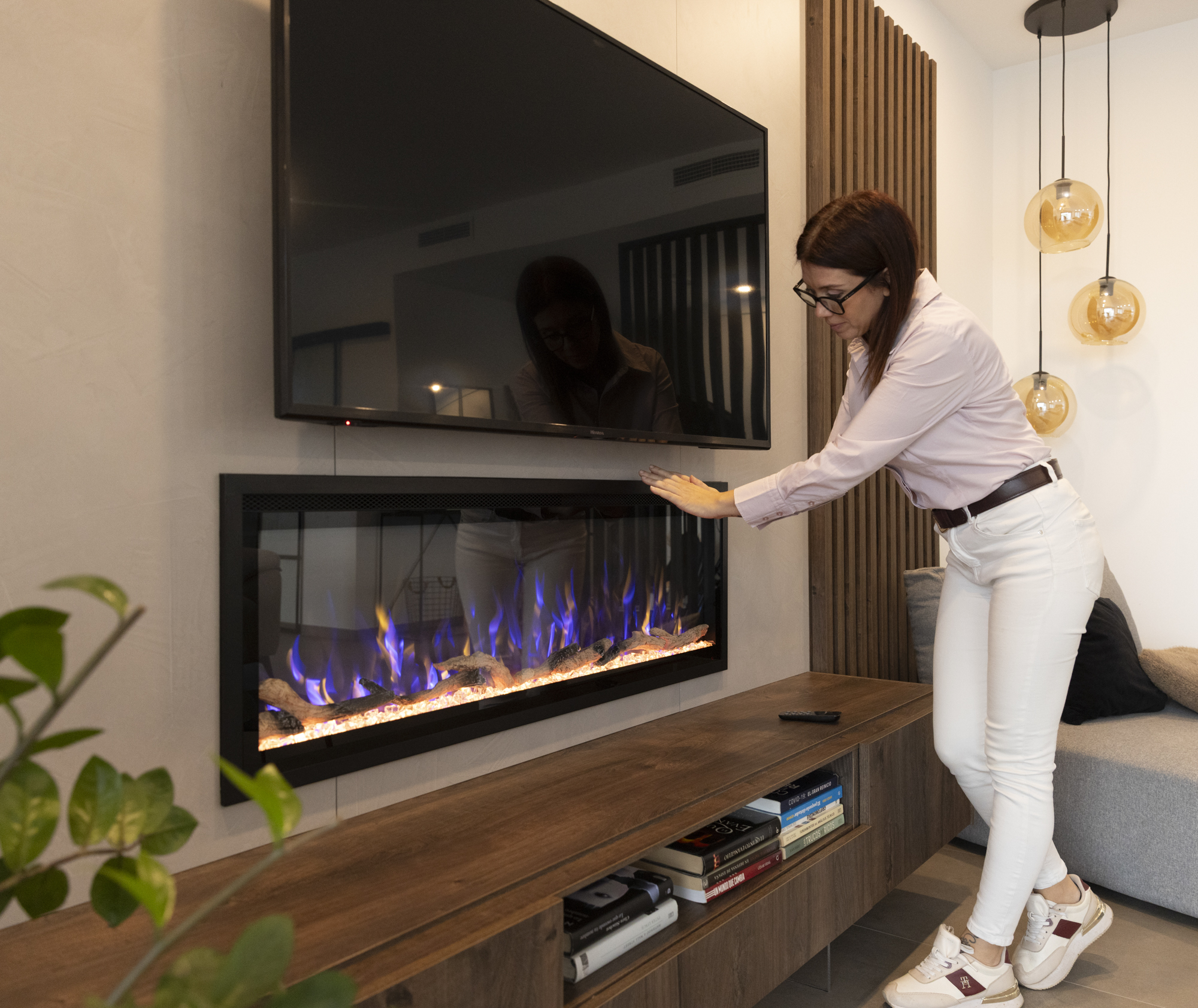 Una mujer toca una chimenea eléctrica debajo de un televisor. La escena incluye un sofá e iluminación decorativa.