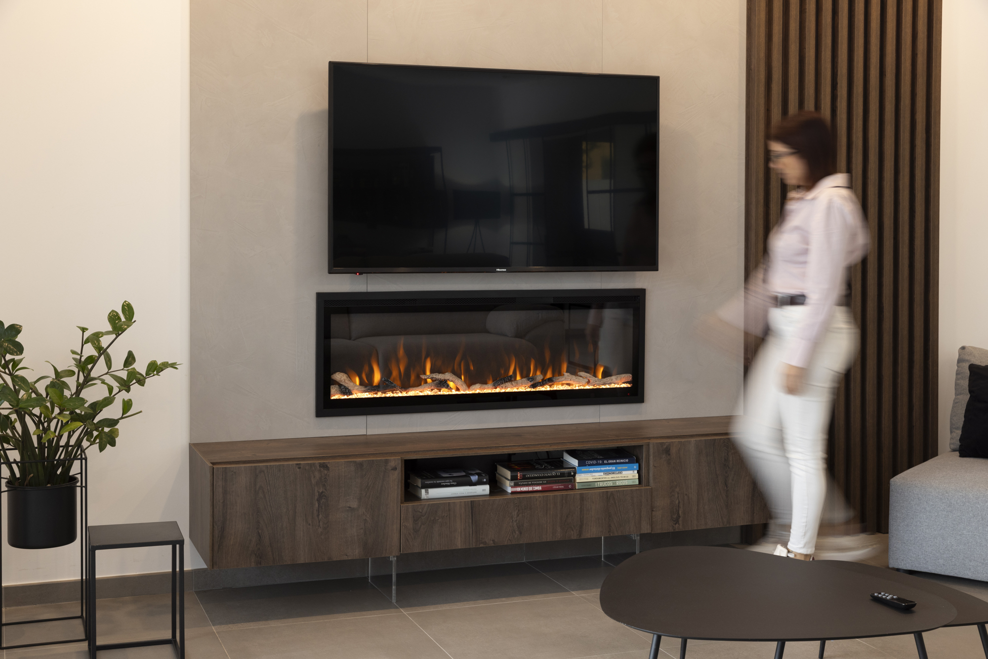 Una sala de estar moderna con TV, chimenea y una persona caminando. Libros en un mueble de madera y una planta a la izquierda.