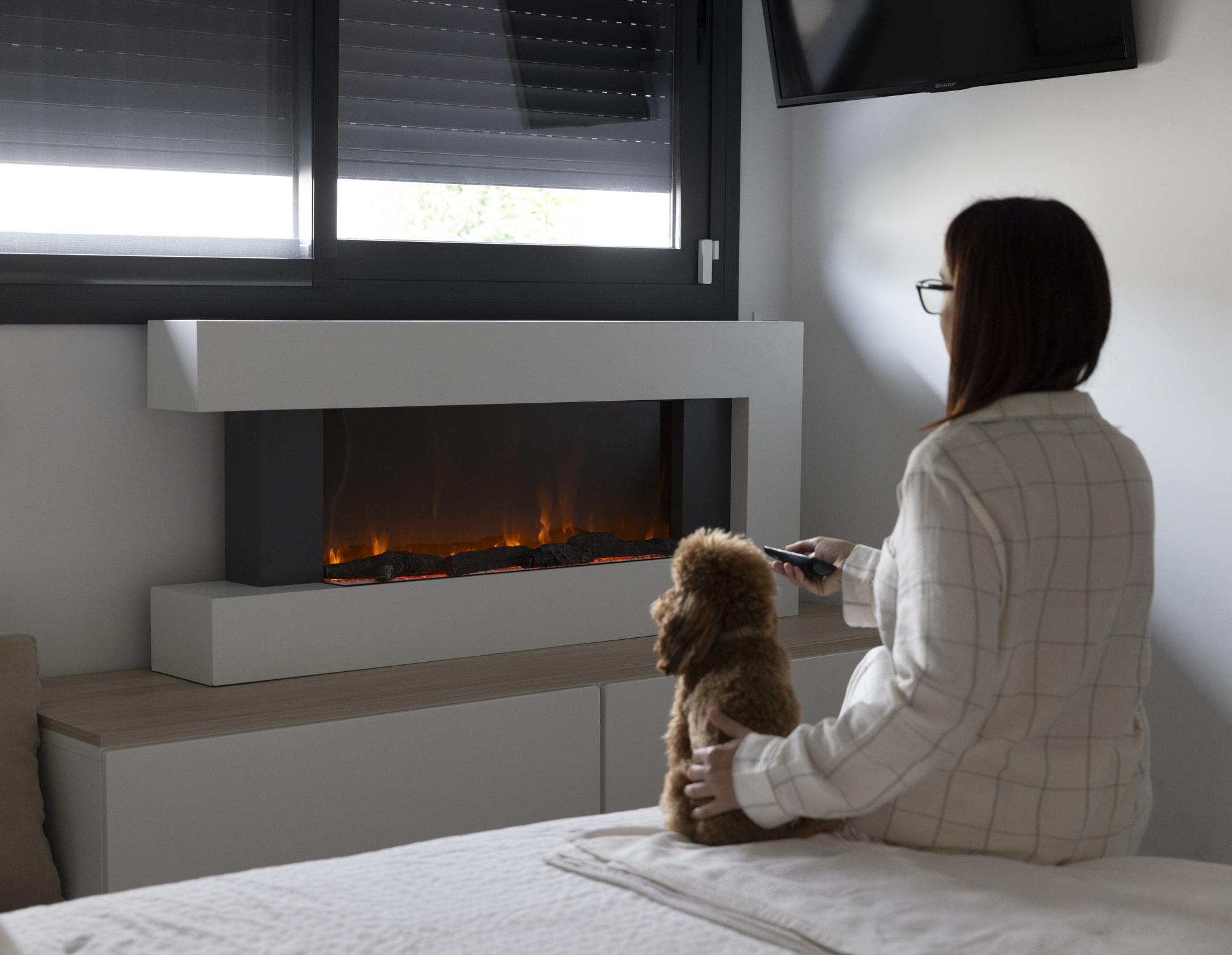 Una mujer con un perro sentada en una cama, usando un control remoto. Una chimenea en el fondo.