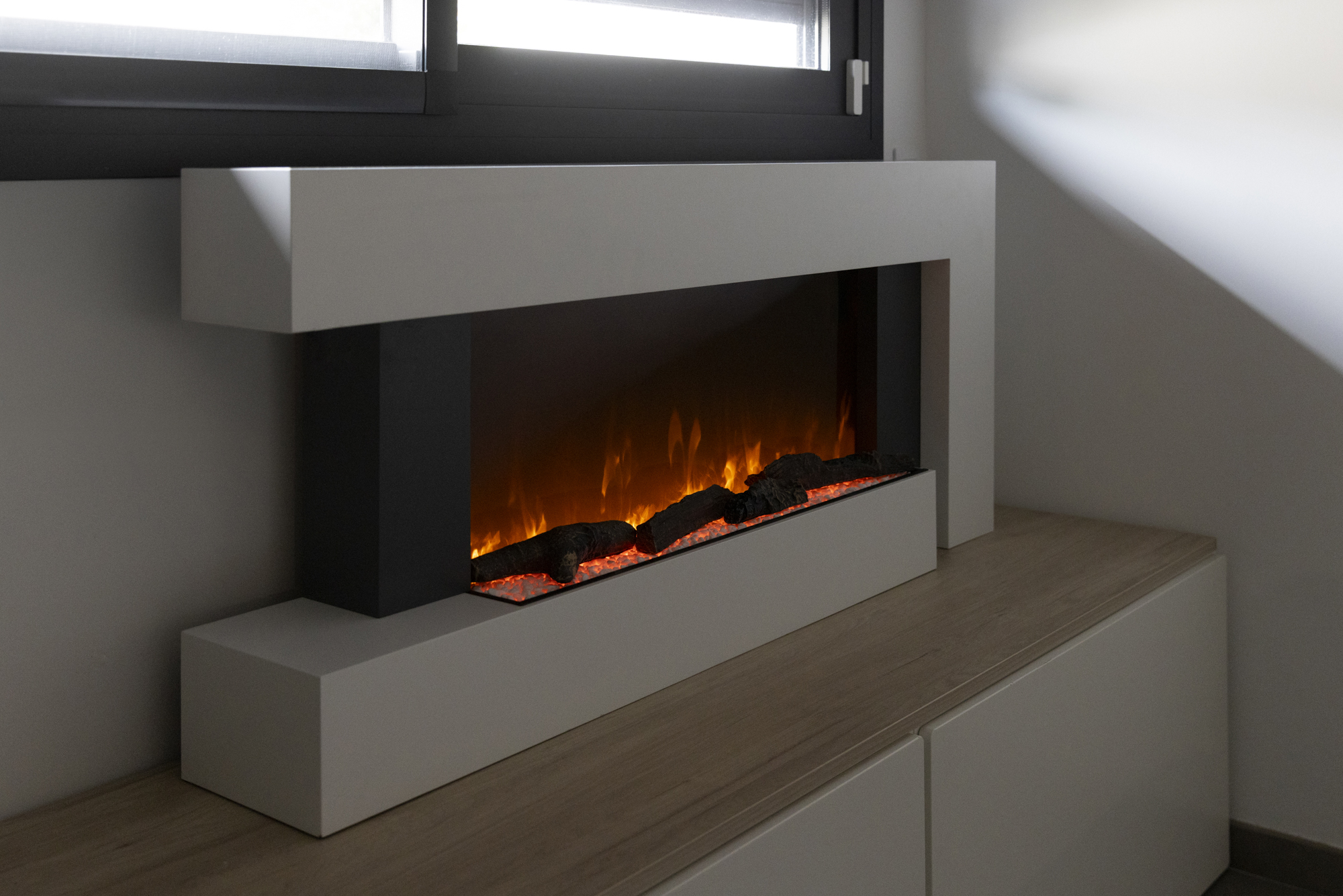 Una chimenea moderna con llamas artificiales. Se encuentra sobre un mueble de madera, con una ventana visible al fondo.