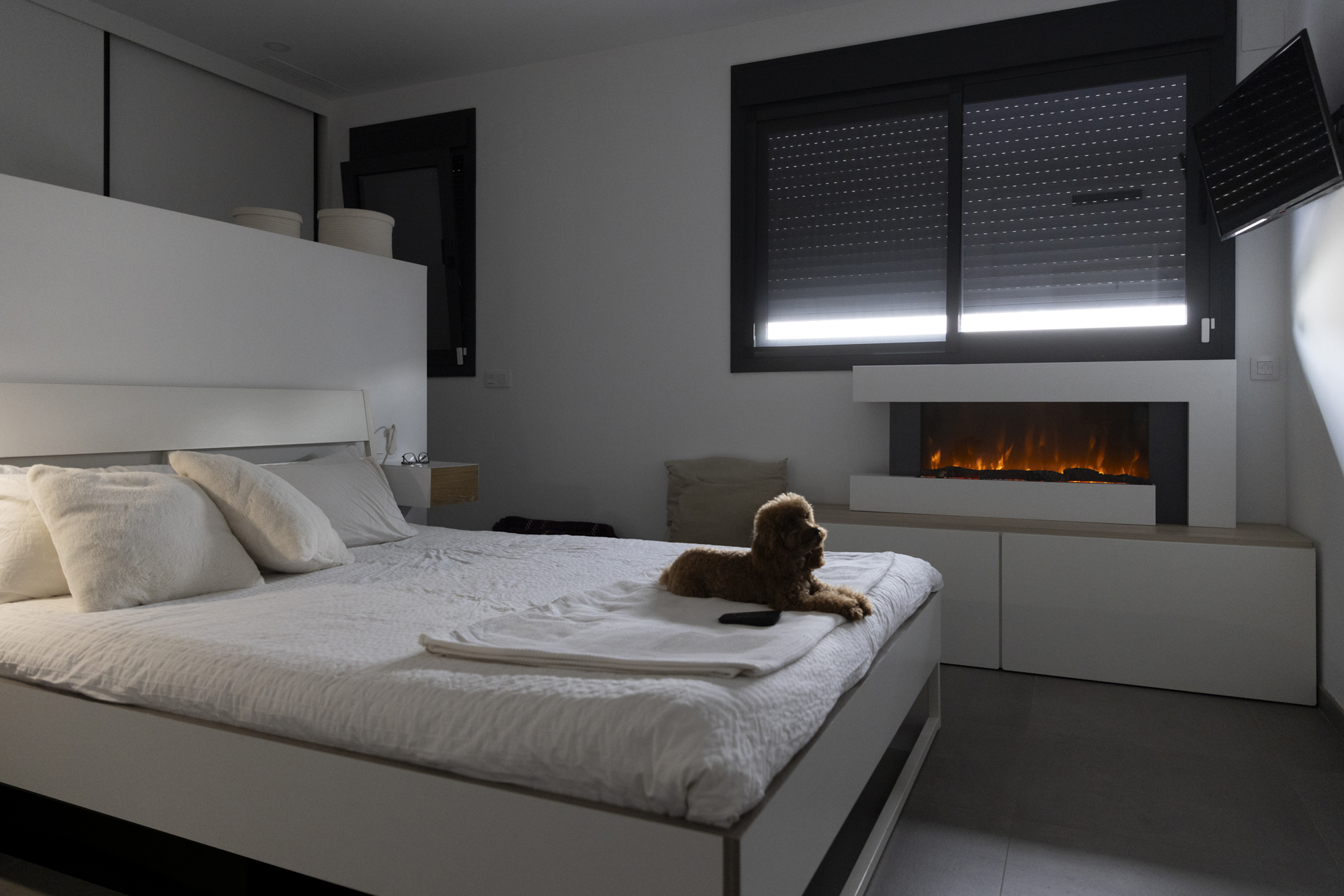Un dormitorio con cama, perro en la cama y chimenea. La habitación está suavemente iluminada, con diseño moderno.