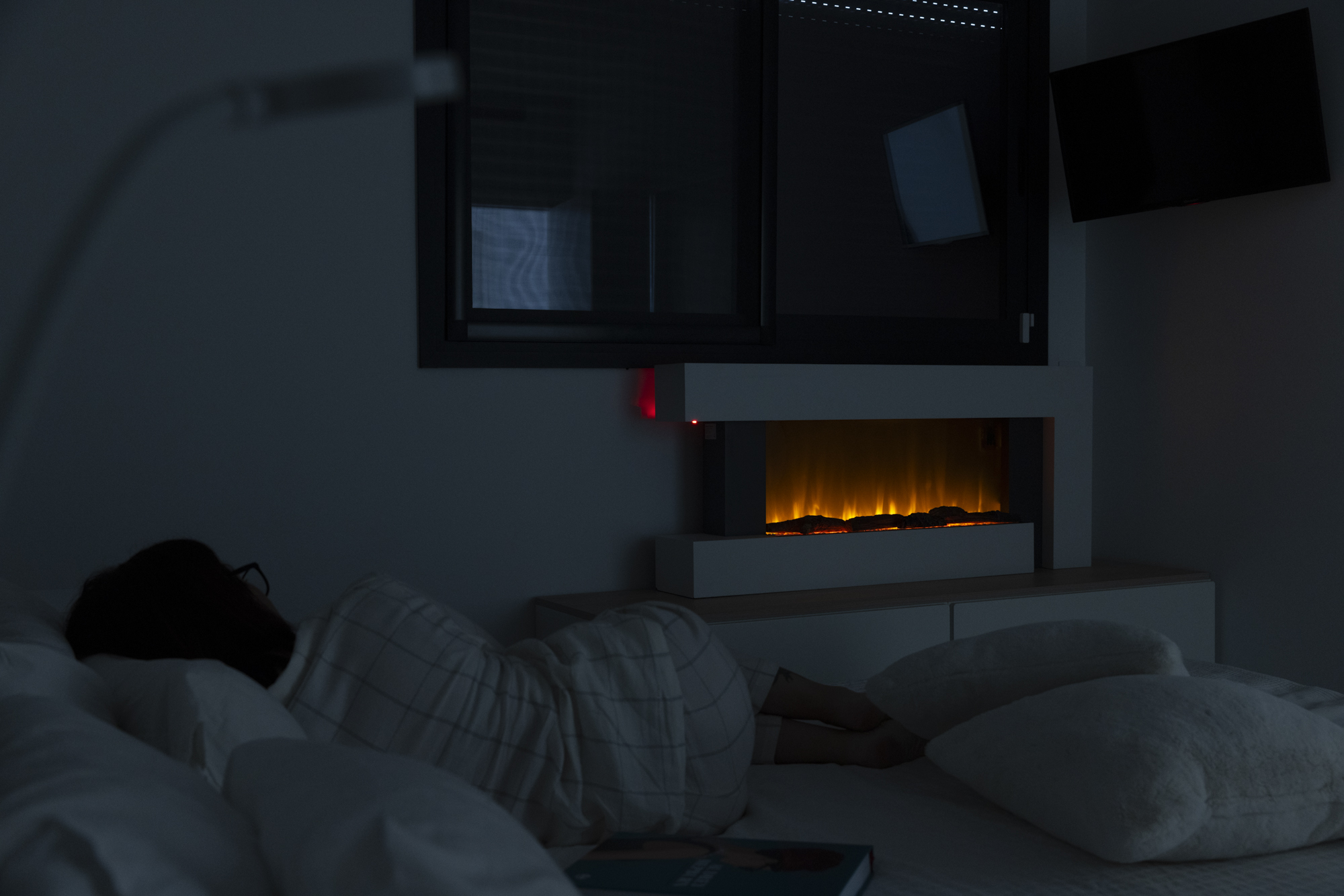 Una persona yace en la cama, una chimenea brilla debajo de un televisor. La iluminación tenue y las sombras dominan la escena. Un libro es visible.