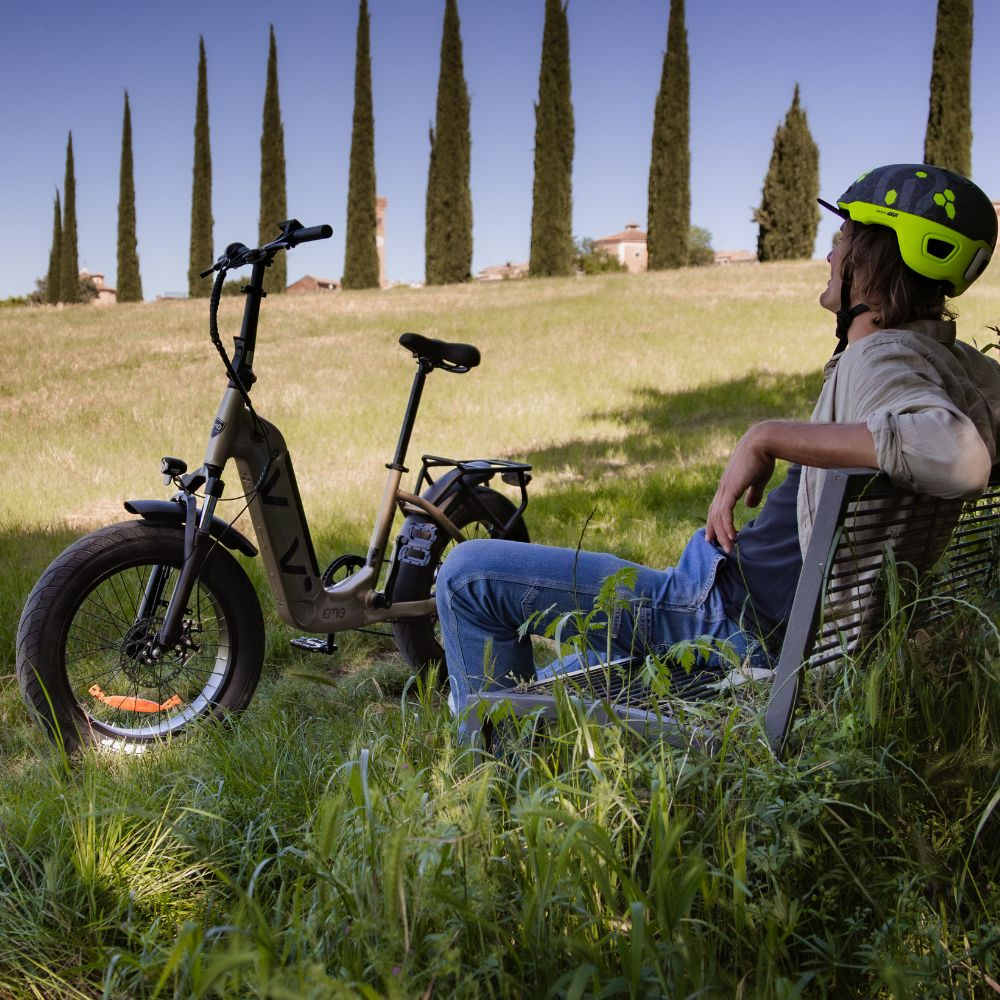 Uomo con casco si riposa su una panchina vicino a una e-bike, campo verde e alberi sullo sfondo sotto un cielo blu.