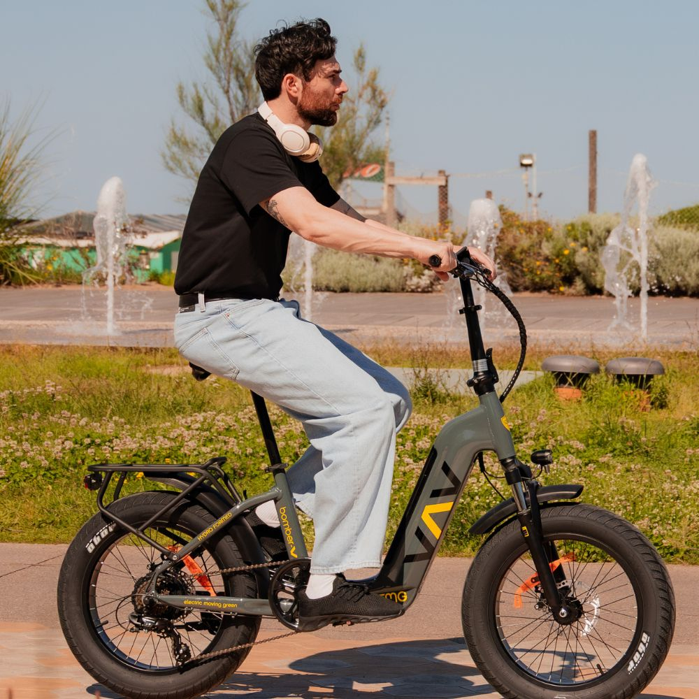 Un uomo guida una bici elettrica in una giornata di sole. Indossa una maglietta nera, jeans e cuffie. In background ci sono fontane e vegetazione.