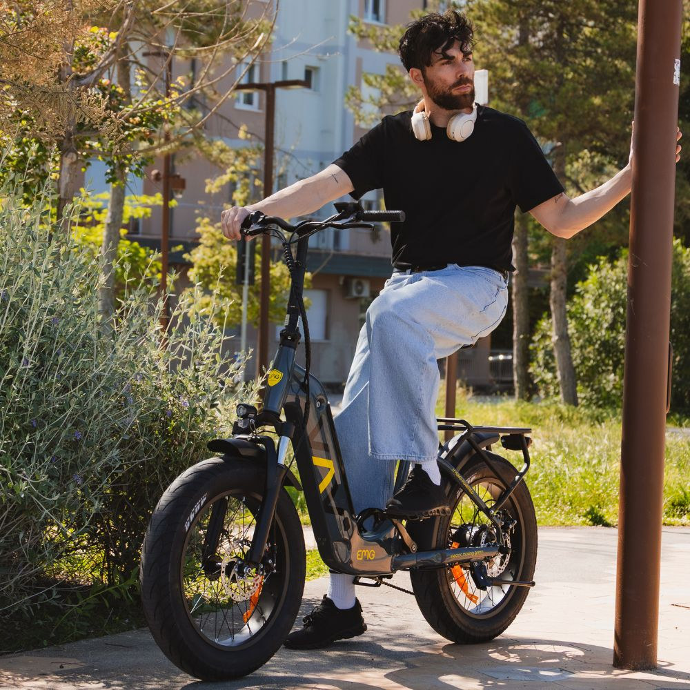 Un uomo seduto su una bici elettrica nera, tiene un palo. Indossa una camicia nera, jeans e cuffie. Alberi ed edifici sullo sfondo.