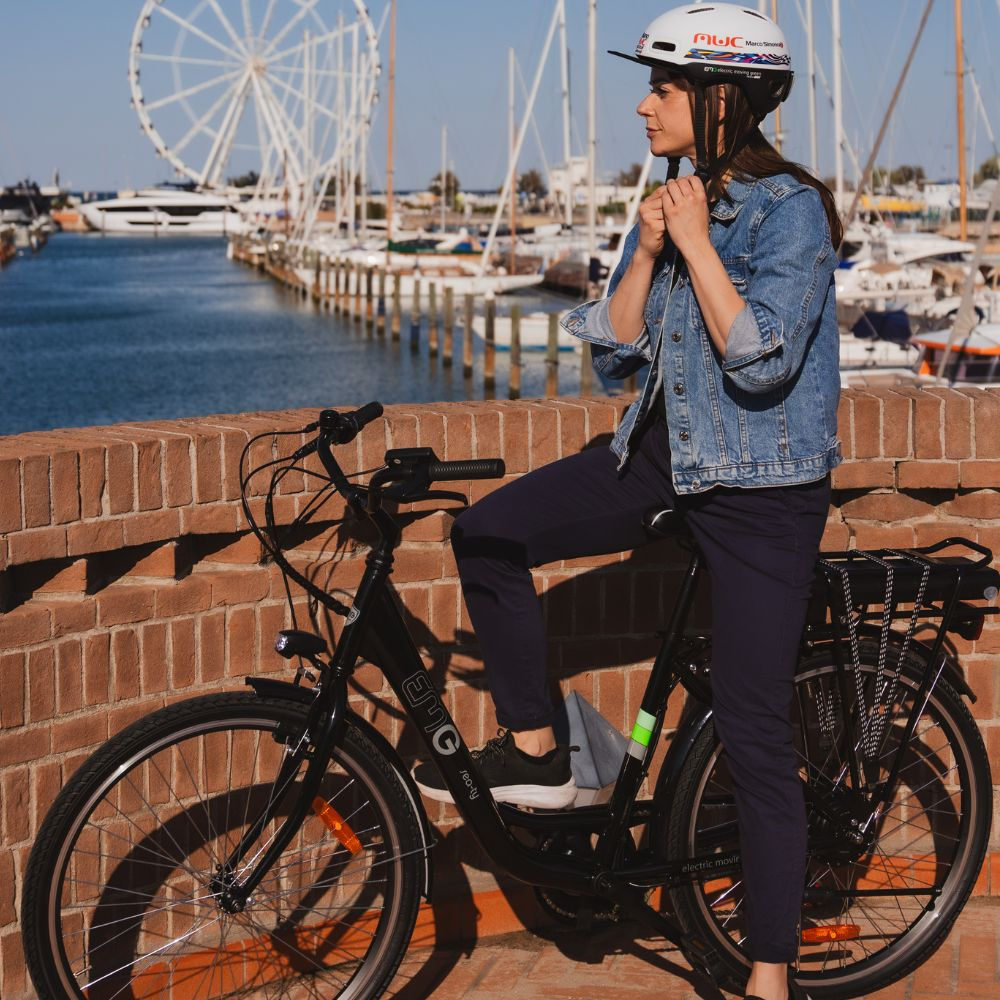 Donna in bicicletta vicino a un muro di mattoni, con casco. Una ruota panoramica e barche sullo sfondo. La bici è nera.
