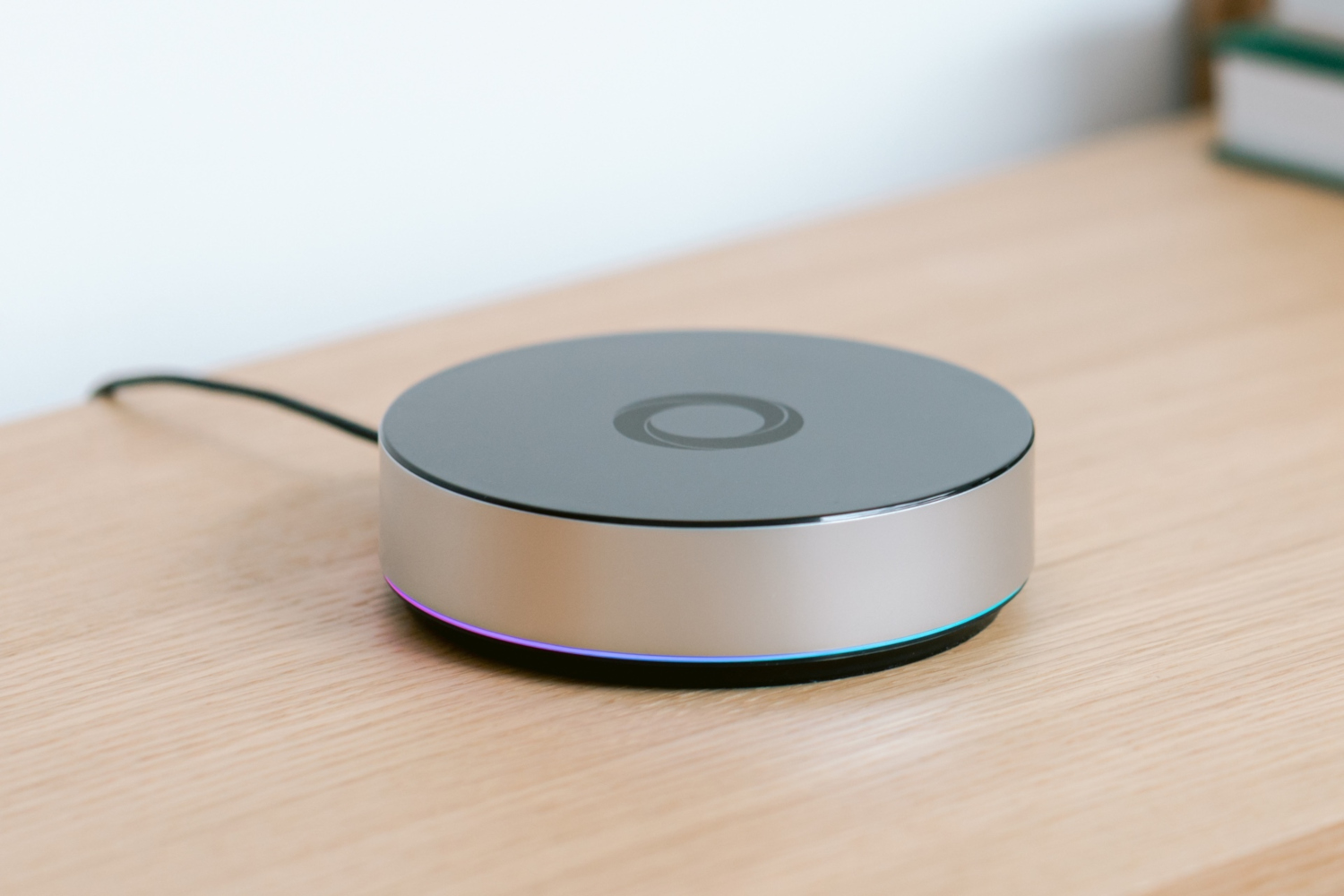 Ein runder, silberner Smart-Home-Hub liegt auf einer Holzoberfläche. Ein dünner, schwarzer Draht geht davon aus. Die Basis leuchtet bunt.