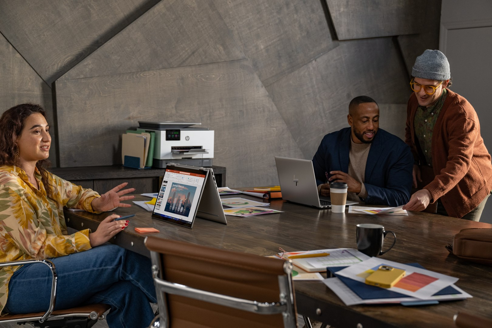 Cztery osoby przy stole z laptopami, drukarką i papierami. Kobieta gestykuluje, dwóch mężczyzn patrzy na papier, a inny pracuje na laptopie.