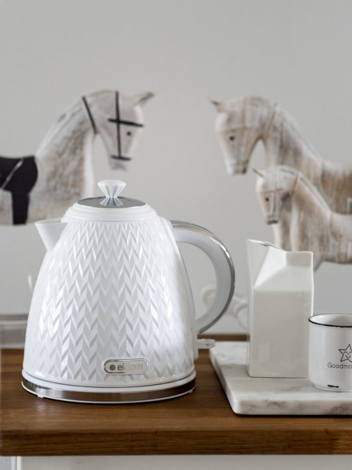 Biały wzorzysty czajnik elektryczny, dzbanek na mleko i kubek na drewnianej powierzchni. Figurki drewnianych koni w tle.