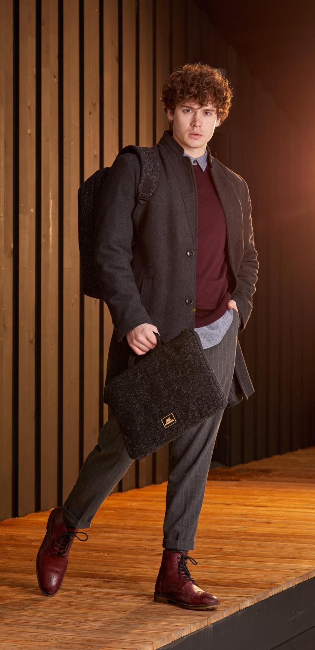 Un uomo in cappotto, maglione e pantaloni, in piedi sul legno, con borsa e zaino, scarpe bordeaux.
