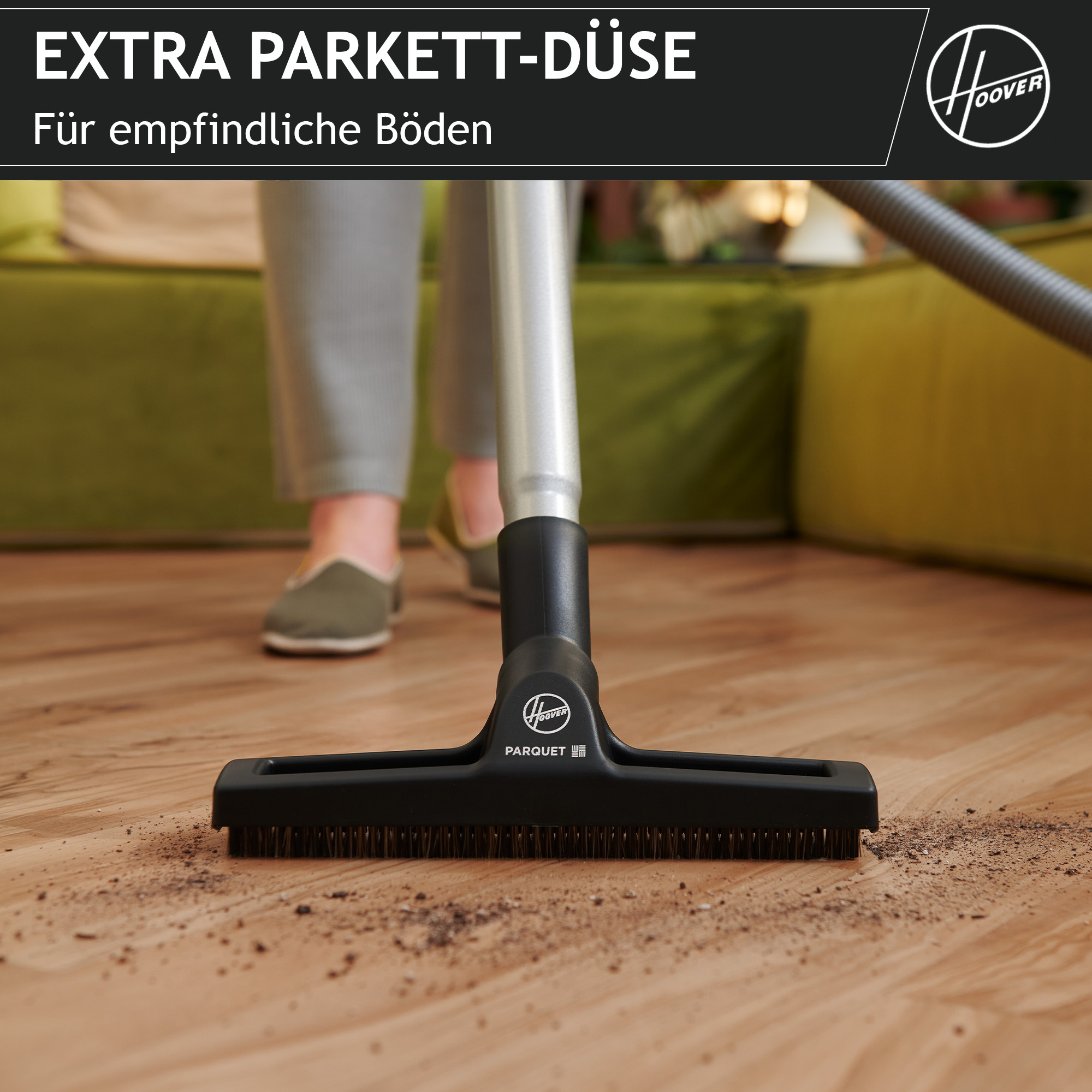 Odkurzacz do parkietu Hoover czyści drewnianą podłogę. W tle nogi osoby i zielona kanapa.