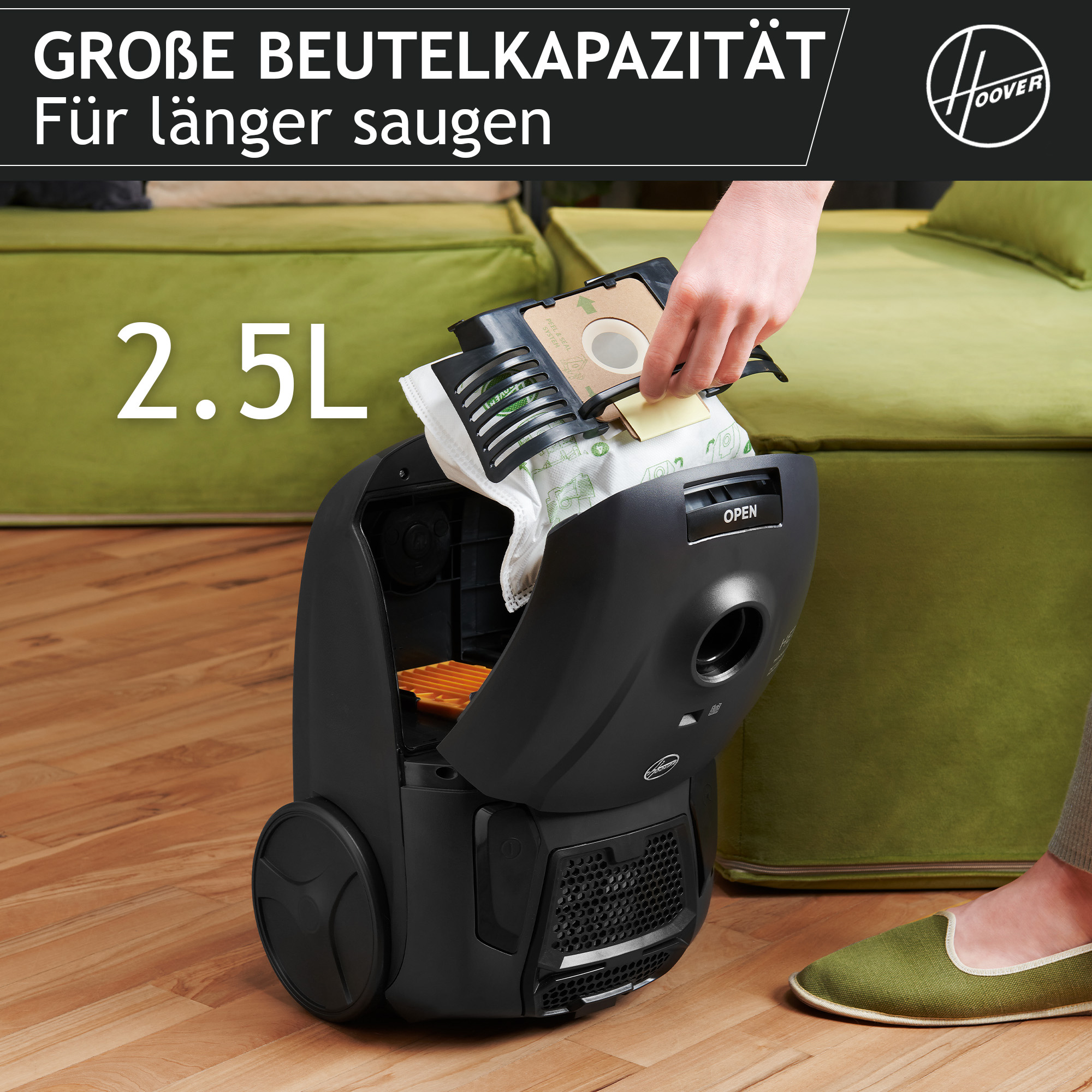 Eine Person ersetzt einen Staubsaugerbeutel in einem schwarzen Hoover-Staubsauger. Im Hintergrund sind grüne Sofas und Holzböden zu sehen.