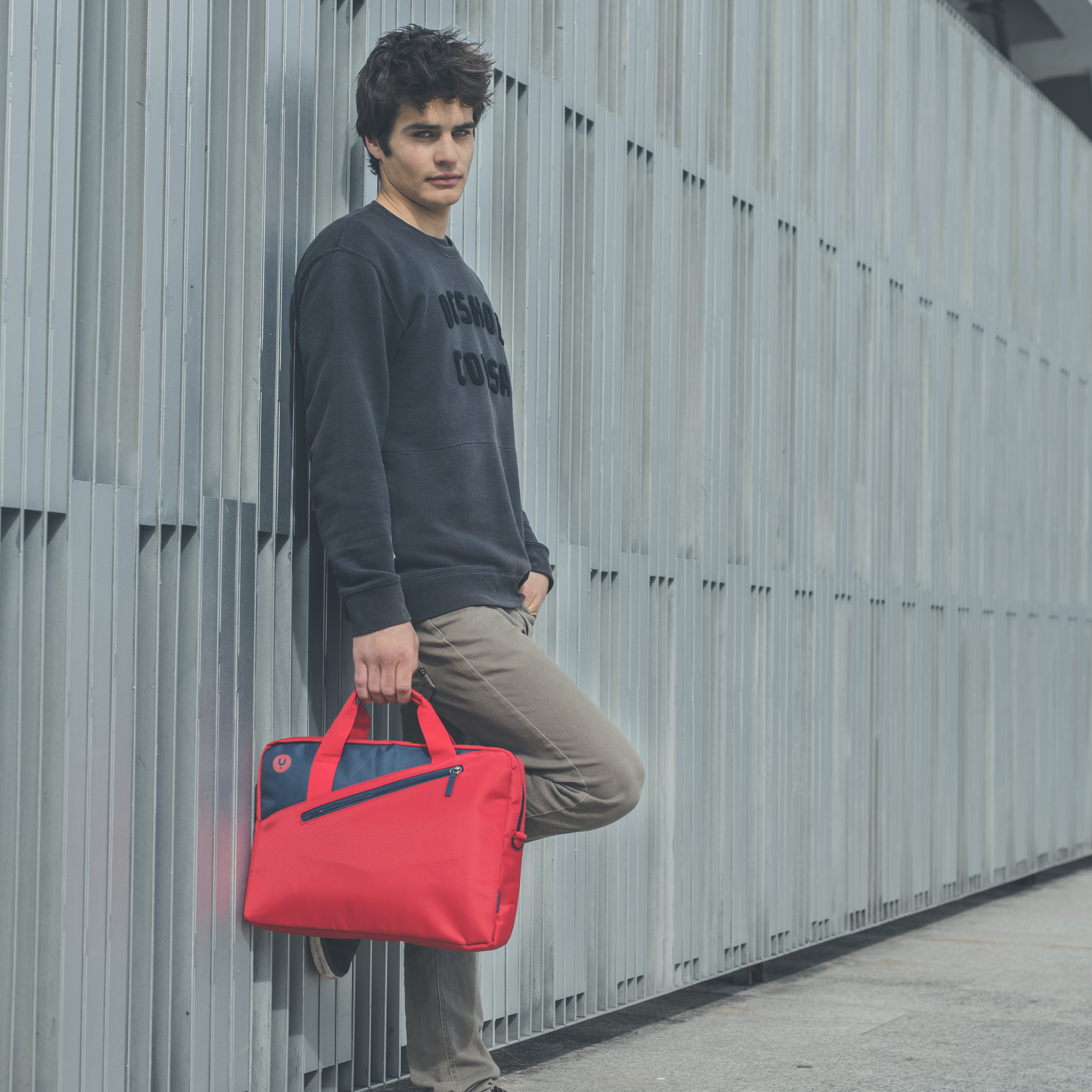 Un hombre se apoya en una pared de metal, sosteniendo una bolsa roja y azul. Viste un suéter oscuro y pantalones caqui.