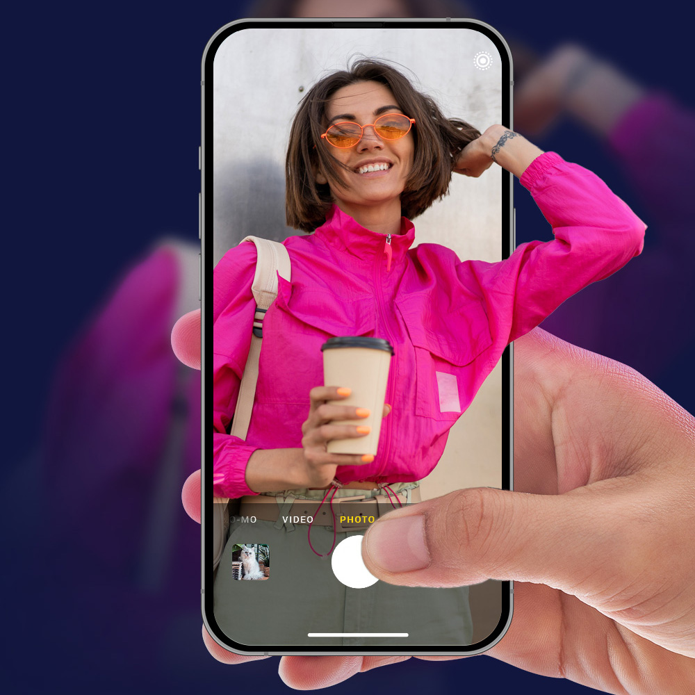 Osoba trzyma telefon, który pokazuje kobietę w różu, pomarańczowych okularach, trzymającą kawę. Pies jest w rogu telefonu.