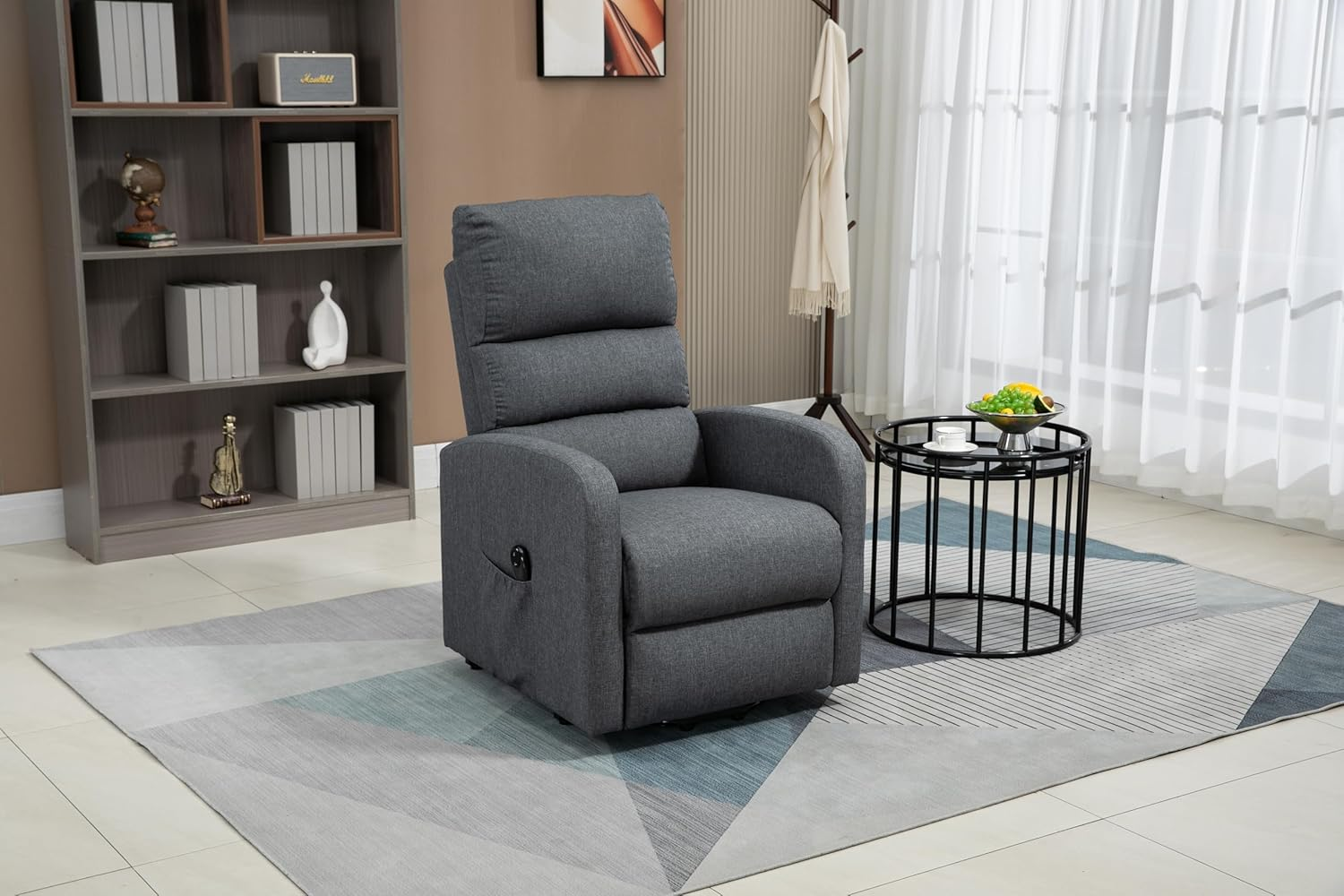 Una silla reclinable gris sobre una alfombra geométrica. Mesa auxiliar con fruta y estantería cerca. Cortinas blancas al fondo.