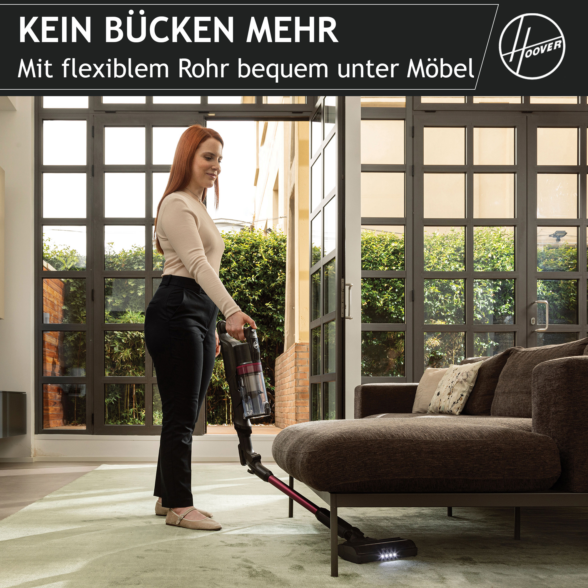Una mujer aspira una habitación con una Hoover. Un sofá marrón a la derecha. Alfombra verde. Grandes ventanas y puertas atrás.