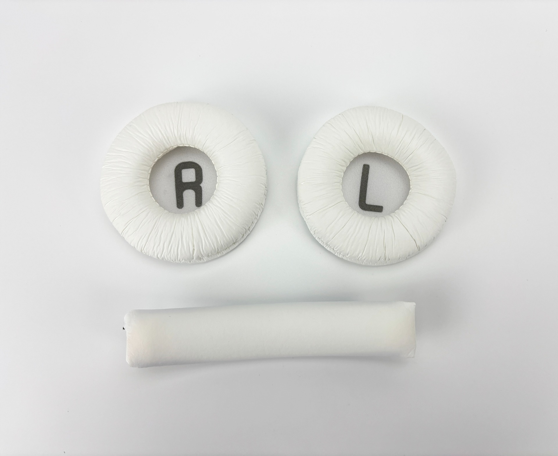Białe części słuchawek na białej powierzchni. Dwie wkładki z 'R' i 'L', a pod nimi podłużna poduszka.