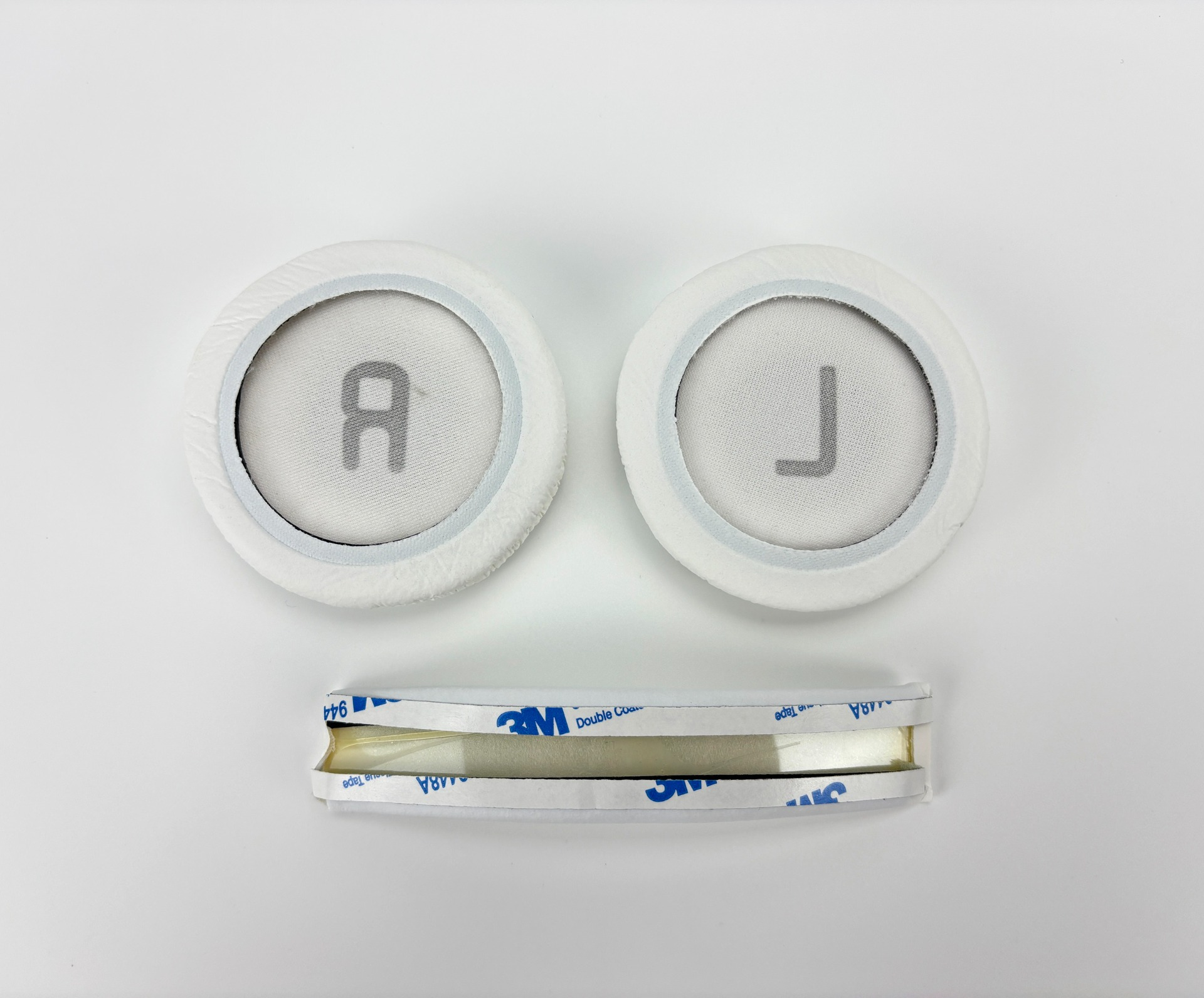 Dwie białe poduszki na uszy z 'R' i 'L', ułożone nad paskiem dwustronnej taśmy klejącej 3M, na białym tle.