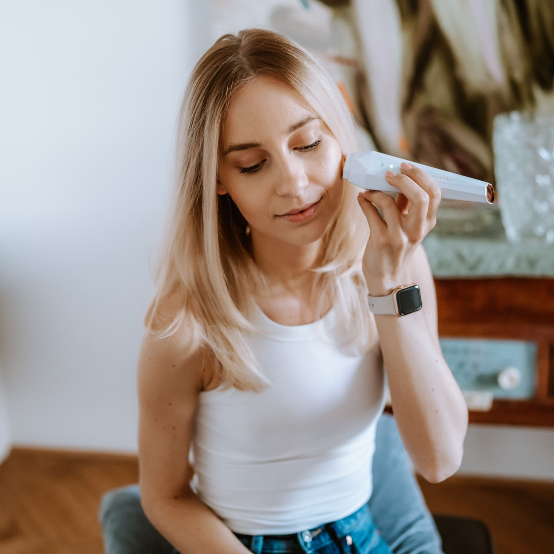 Eine Frau benutzt ein Hautpflegegerät. Sie hat blondes Haar und trägt ein weißes Tanktop. An ihrem Handgelenk ist eine Uhr.