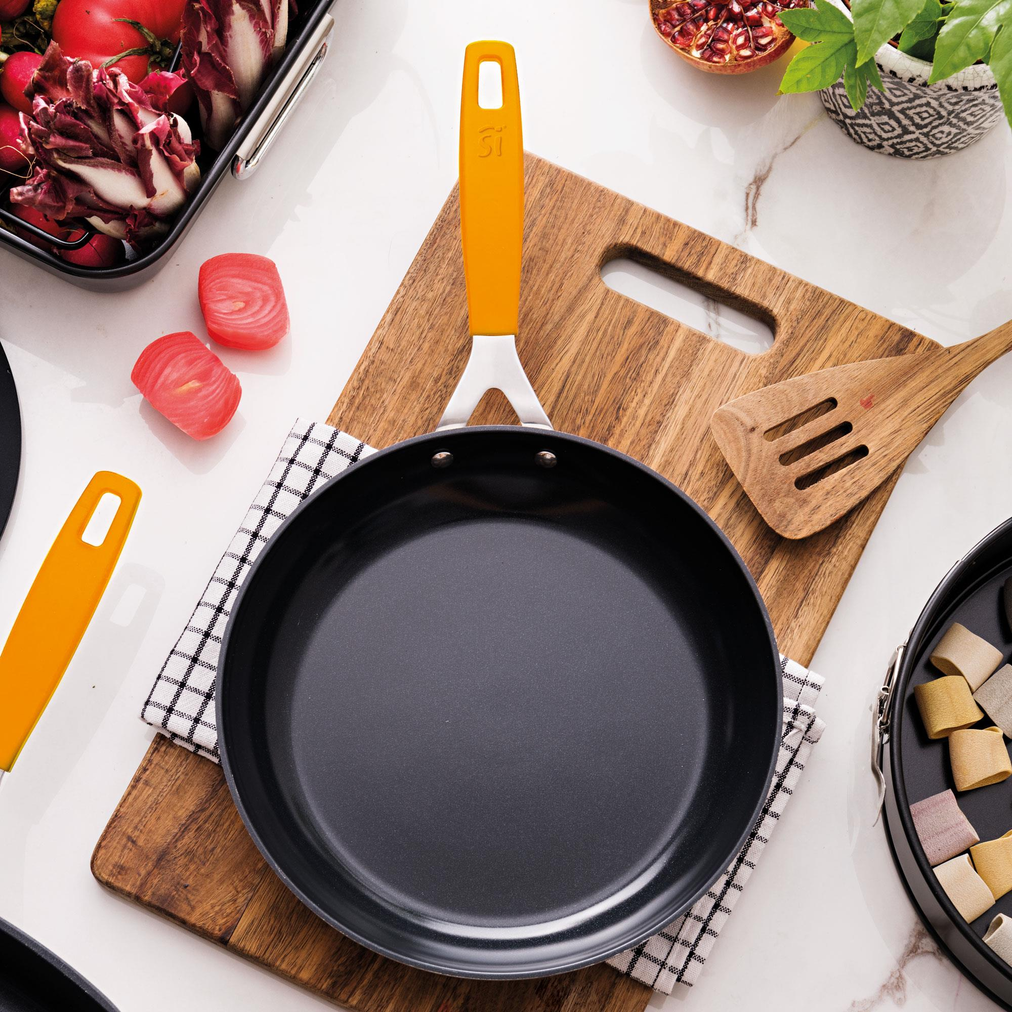 Una sartén con mango naranja descansa sobre una tabla de madera con verduras y otros utensilios de cocina.