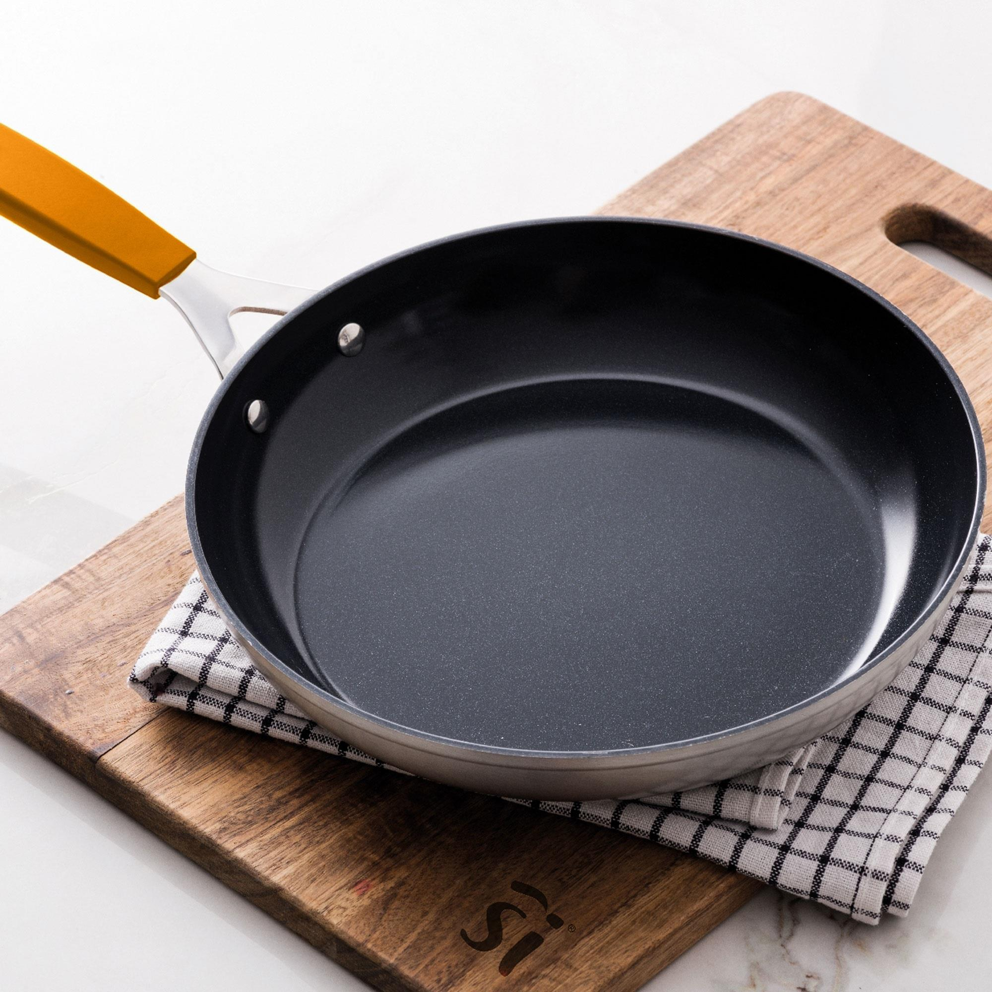 Una sartén con mango naranja descansa sobre una tabla de madera con un paño a cuadros. Fondo blanco.