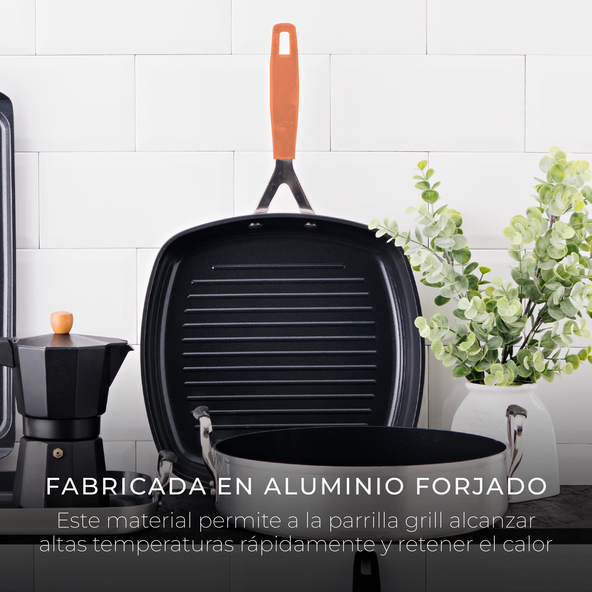 Una sartén de parrilla negra con mango naranja, una cafetera negra y un jarrón con plantas verdes sobre baldosas blancas.
