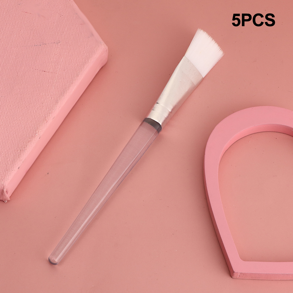 Ein Kosmetikpinsel mit transparentem Griff und weißen Borsten liegt auf einer rosa Oberfläche. Eine rosa Form und ein rosa Block sind ebenfalls vorhanden.