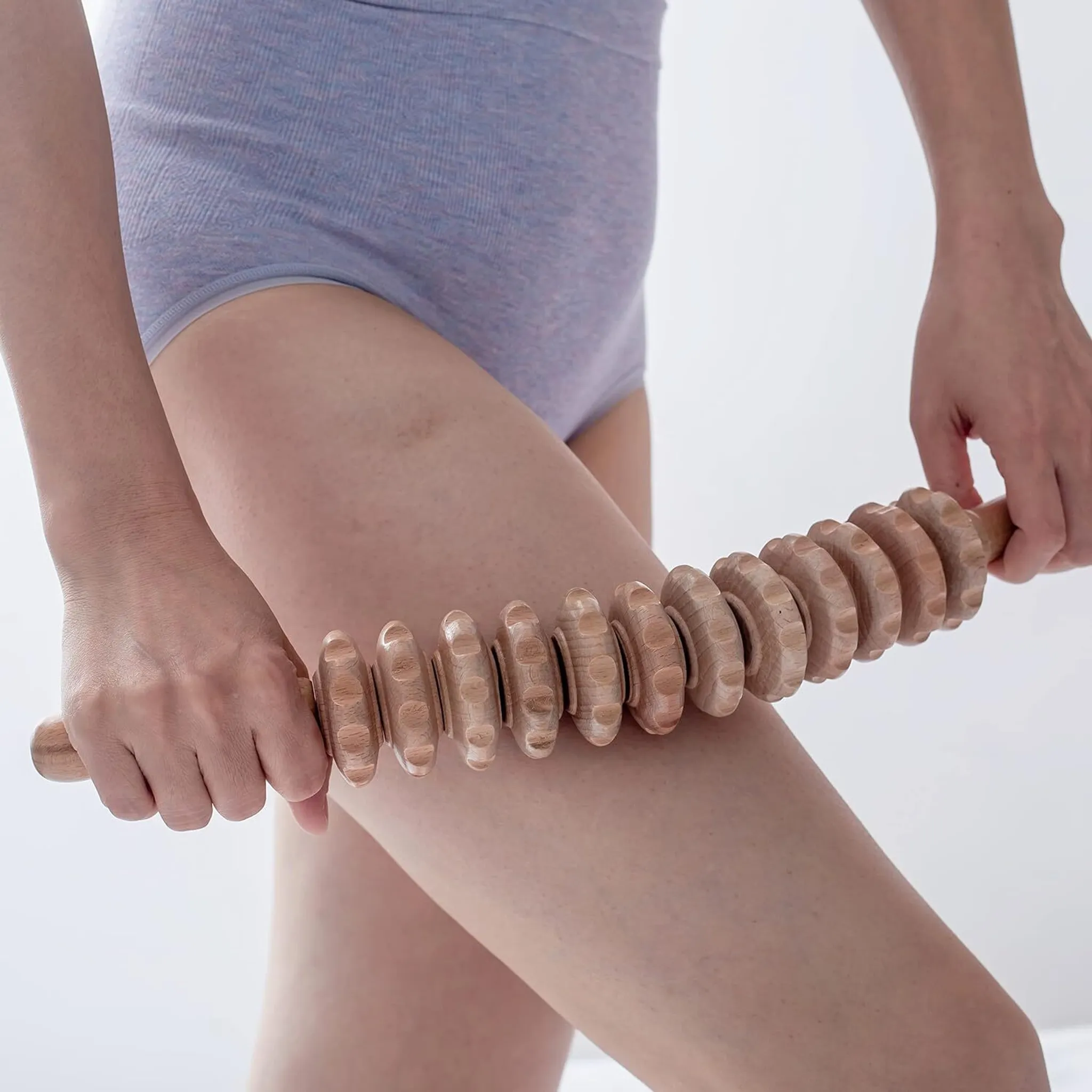 Eine Person benutzt eine hölzerne Massagerolle an ihrem Oberschenkel. Sie trägt graue Unterwäsche, der Hintergrund ist weiß.