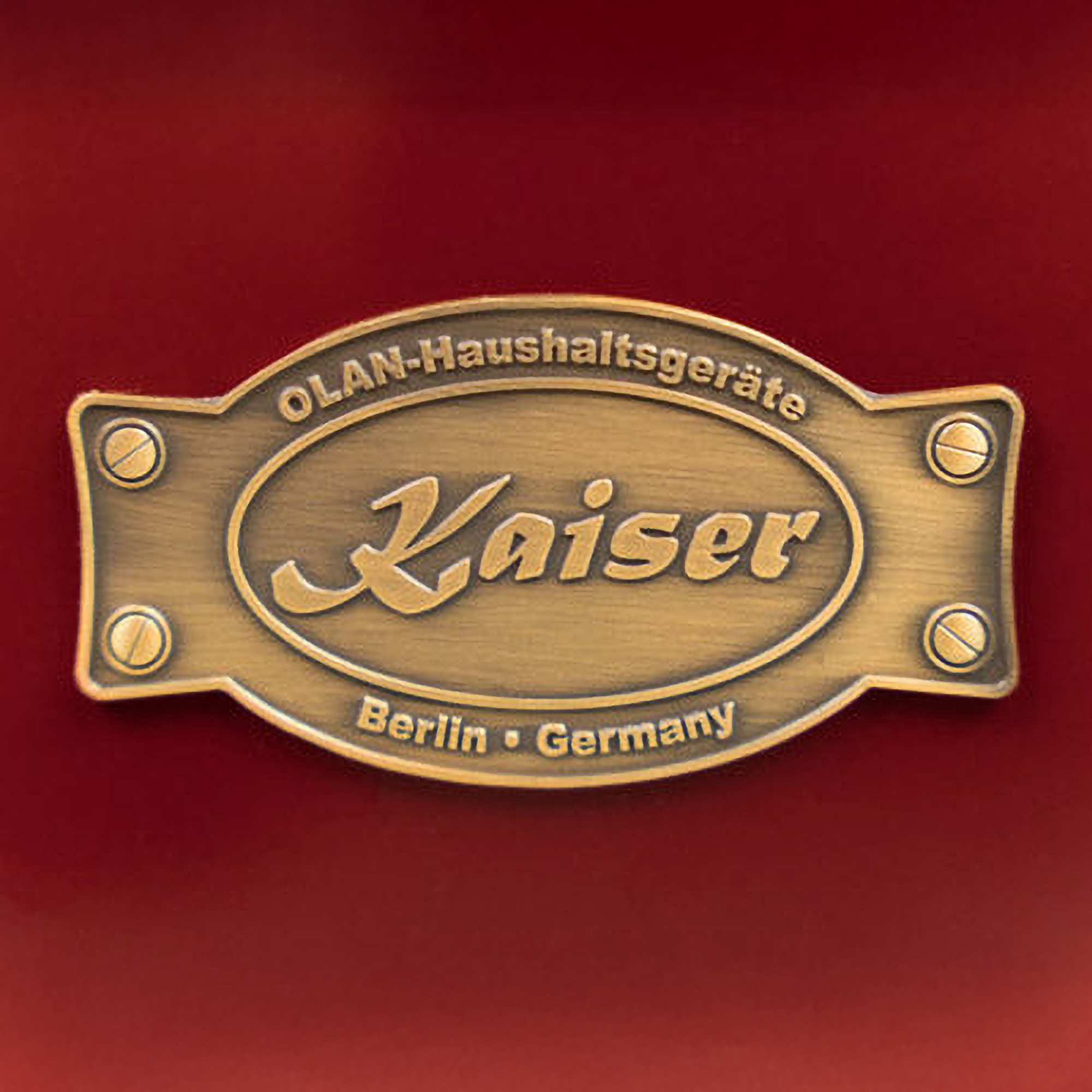 Ein Messing-Kaiser-Emblem auf rotem Hintergrund. Es lautet 'OLAN-Haushaltgeräte Kaiser Berlin Germany'.