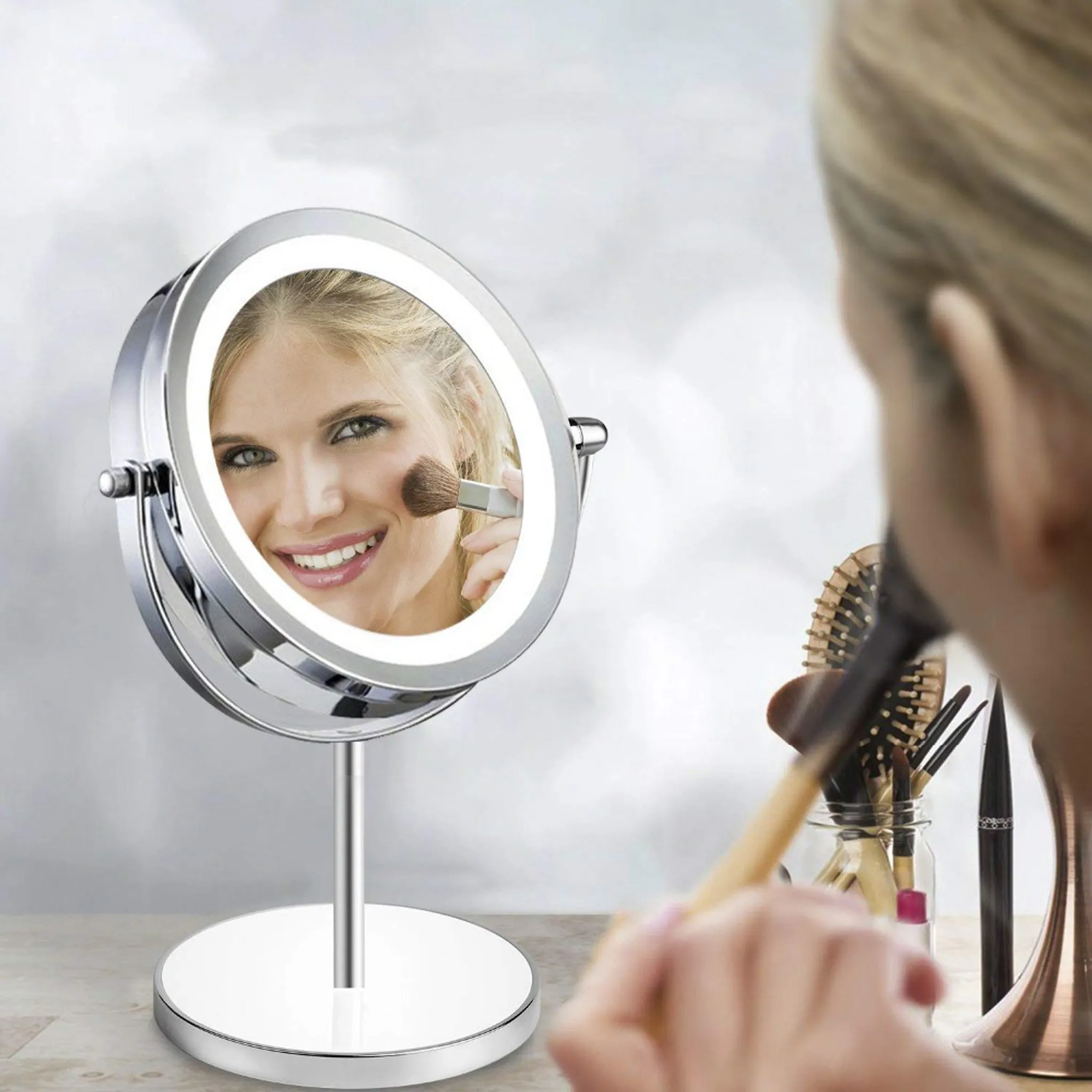 Eine Frau trägt Make-up auf. Ein Spiegel zeigt ihr lächelndes Gesicht. Pinsel und Kosmetika stehen auf dem Tisch.
