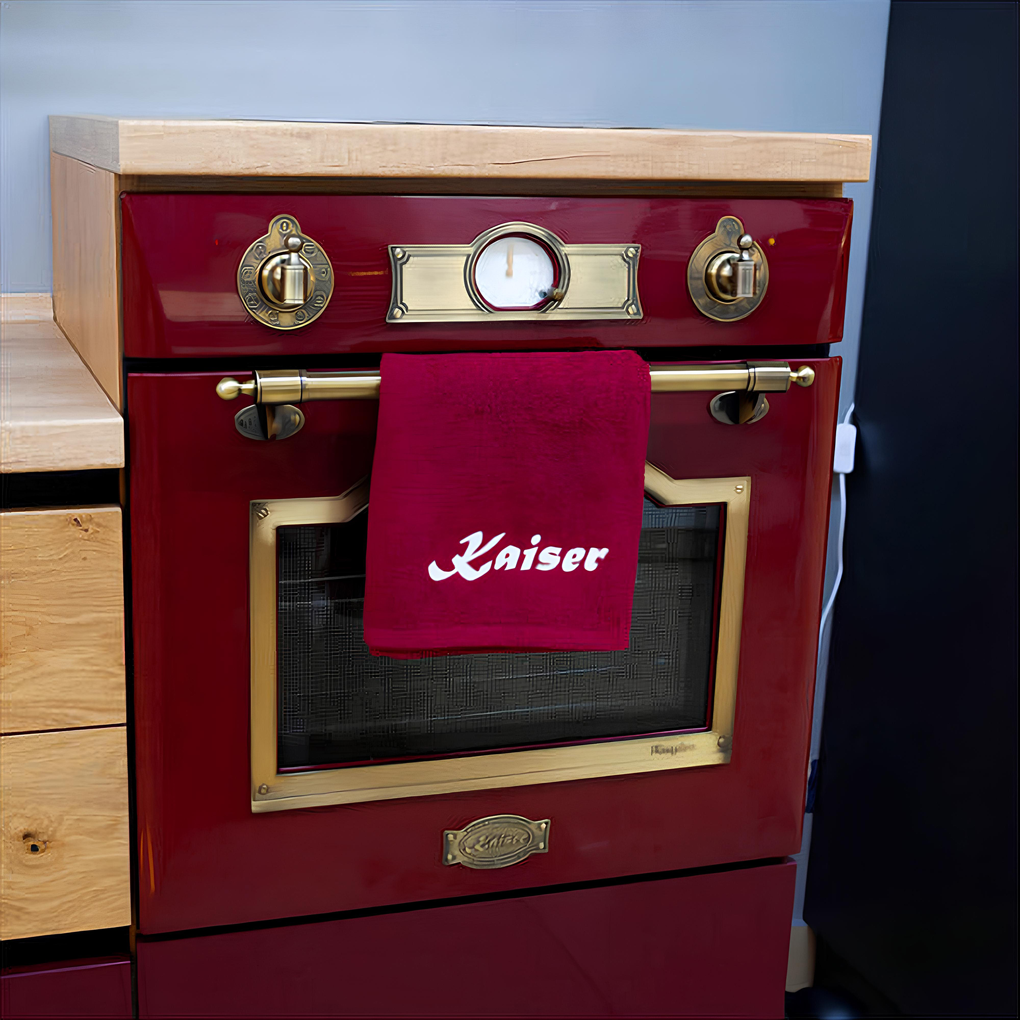 Ein roter Ofen mit goldfarbenen Verzierungen. Ein Handtuch mit dem Namen „Kaiser“ hängt vorne. Oben befindet sich eine Holzplatte.