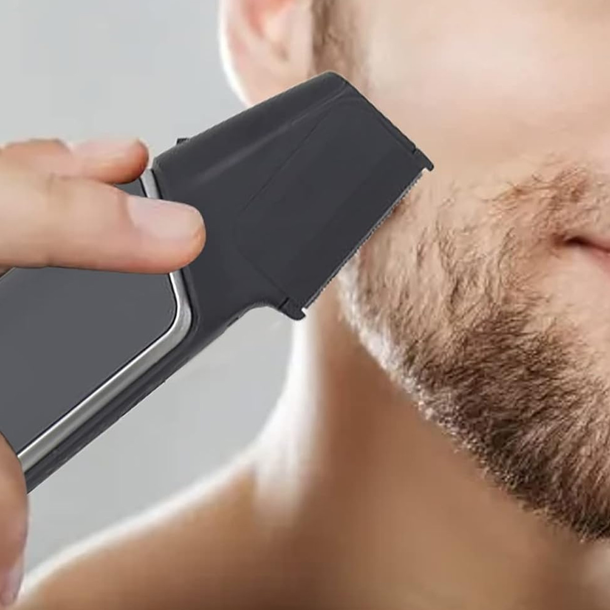 Ein Mann rasiert seinen Bart mit einem elektrischen Trimmer. Der Trimmer ist schwarz und wird in der Nähe seines Gesichts gehalten, der Hintergrund ist verschwommen.