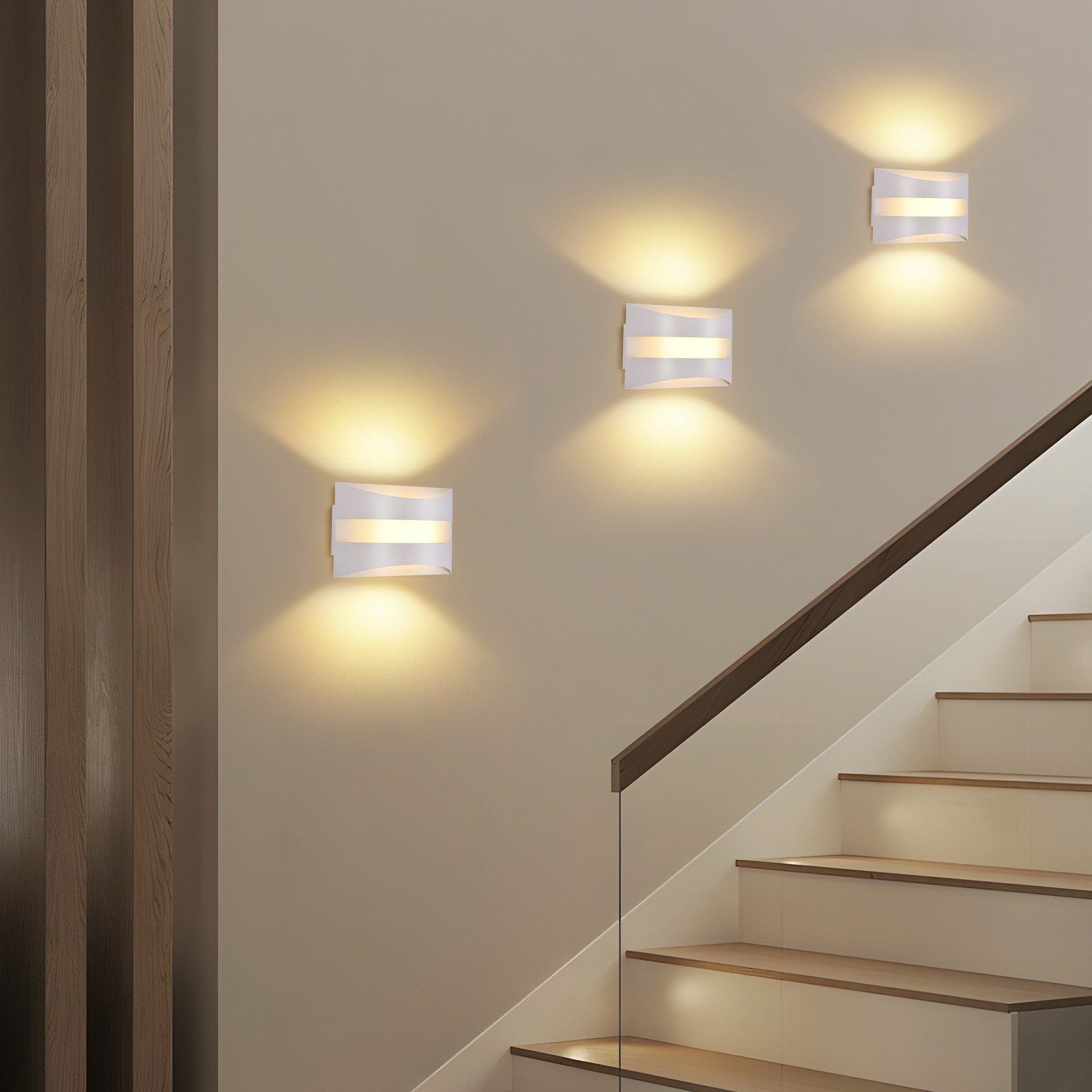 Lampade da parete bianche illuminano una scala. Scala in legno con corrimano marrone. Parete beige con pannelli in legno a sinistra.